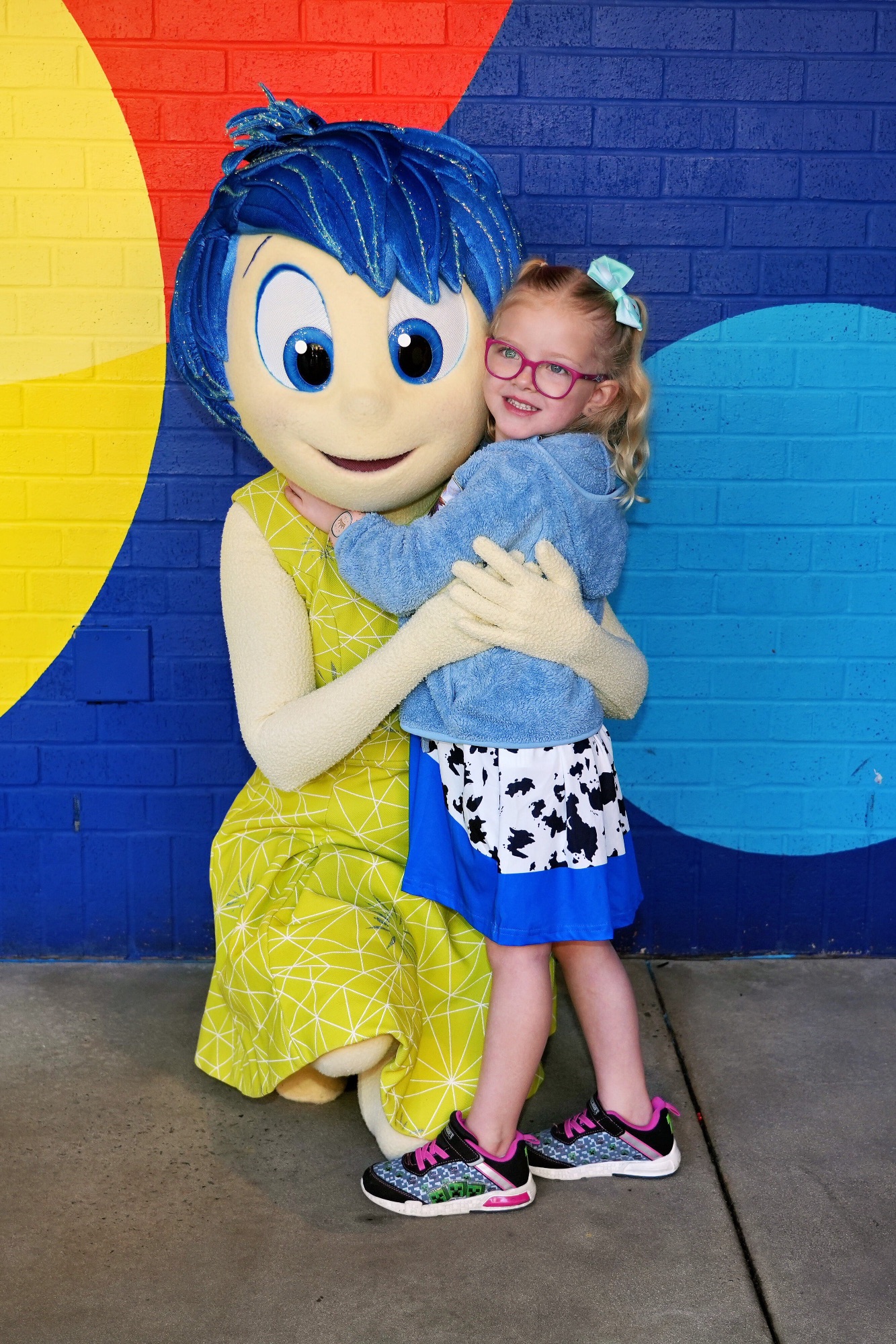 Woman and young child posing with a character in blue outfit at an indoor attraction with colorful backdrop