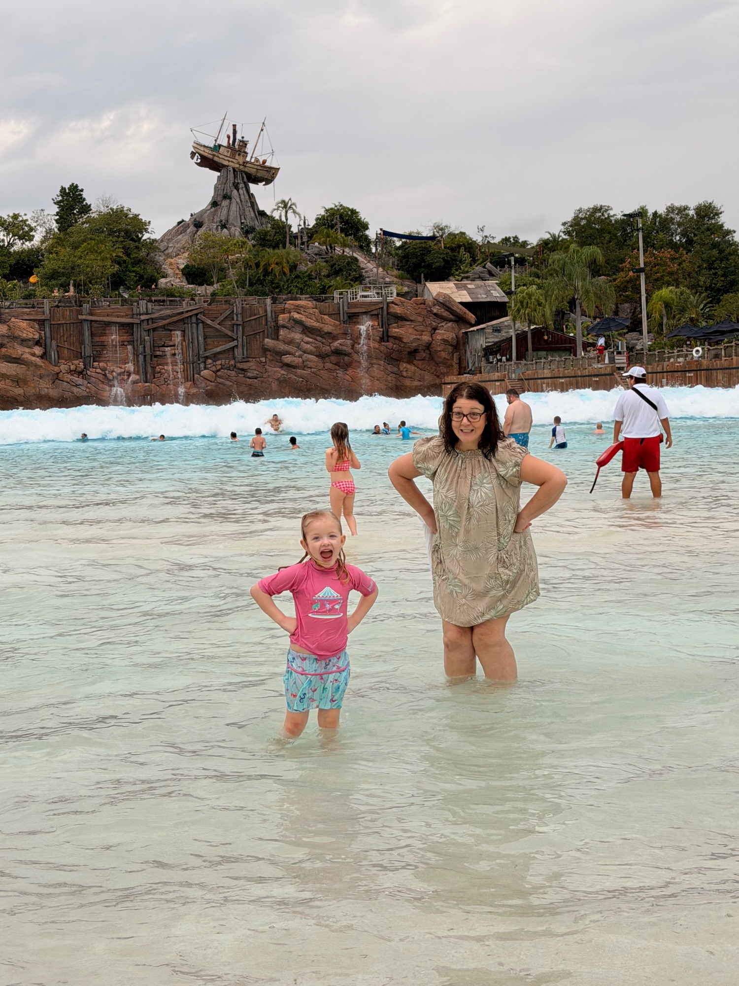 Woman and young child wading in shallow turquoise water at a beach with a large ship structure visible in background