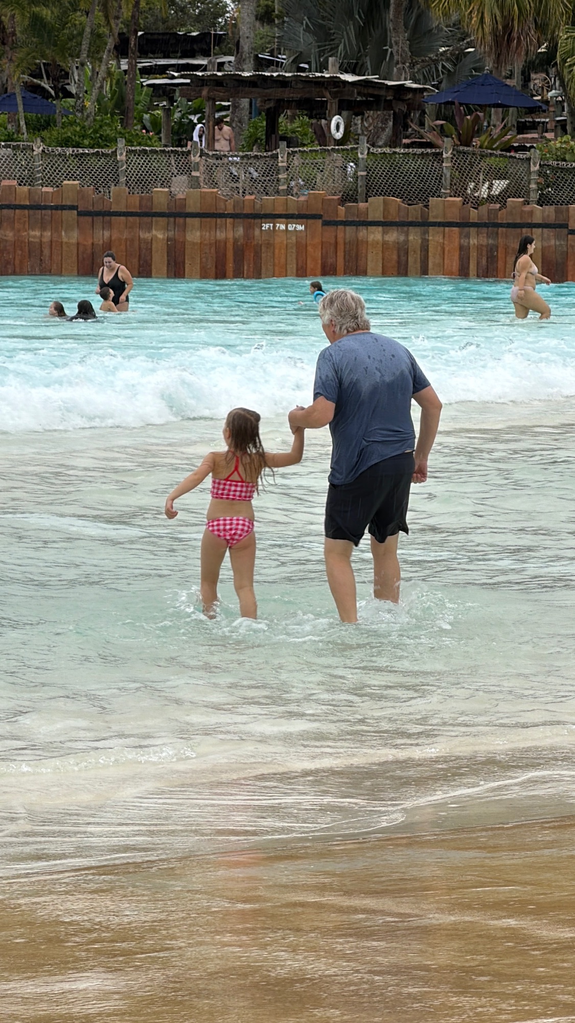 Older man and young girl in pink swimsuit entering the ocean together at a beach with wooden structures behind them