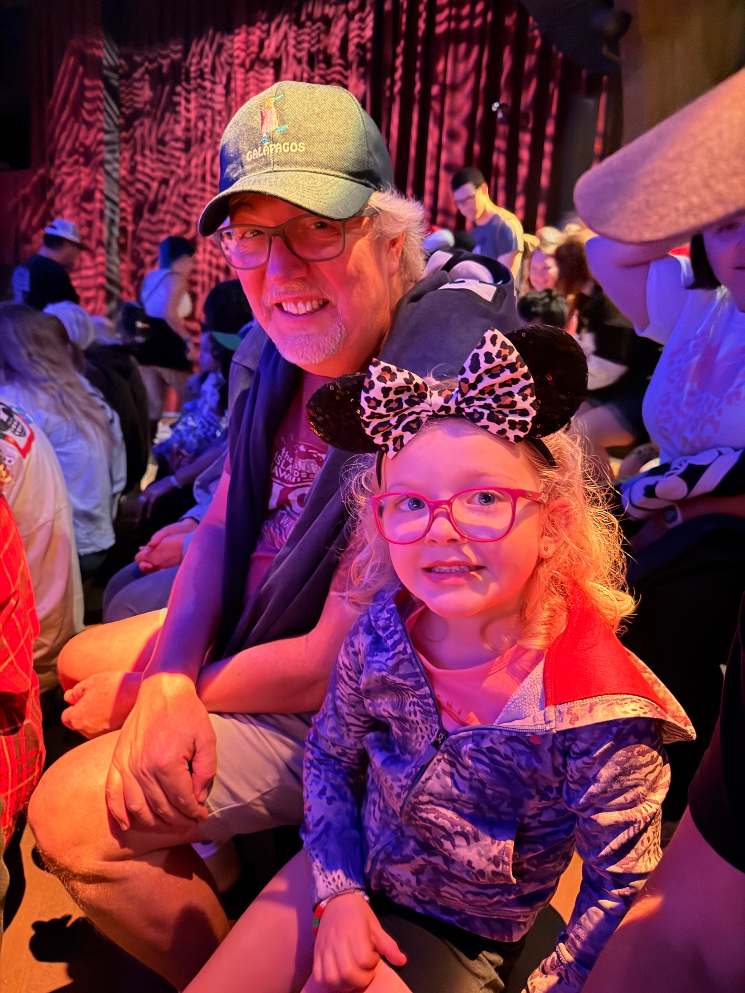 Older man wearing a Galapagos cap posing with a young girl in colorful costume at an indoor entertainment venue