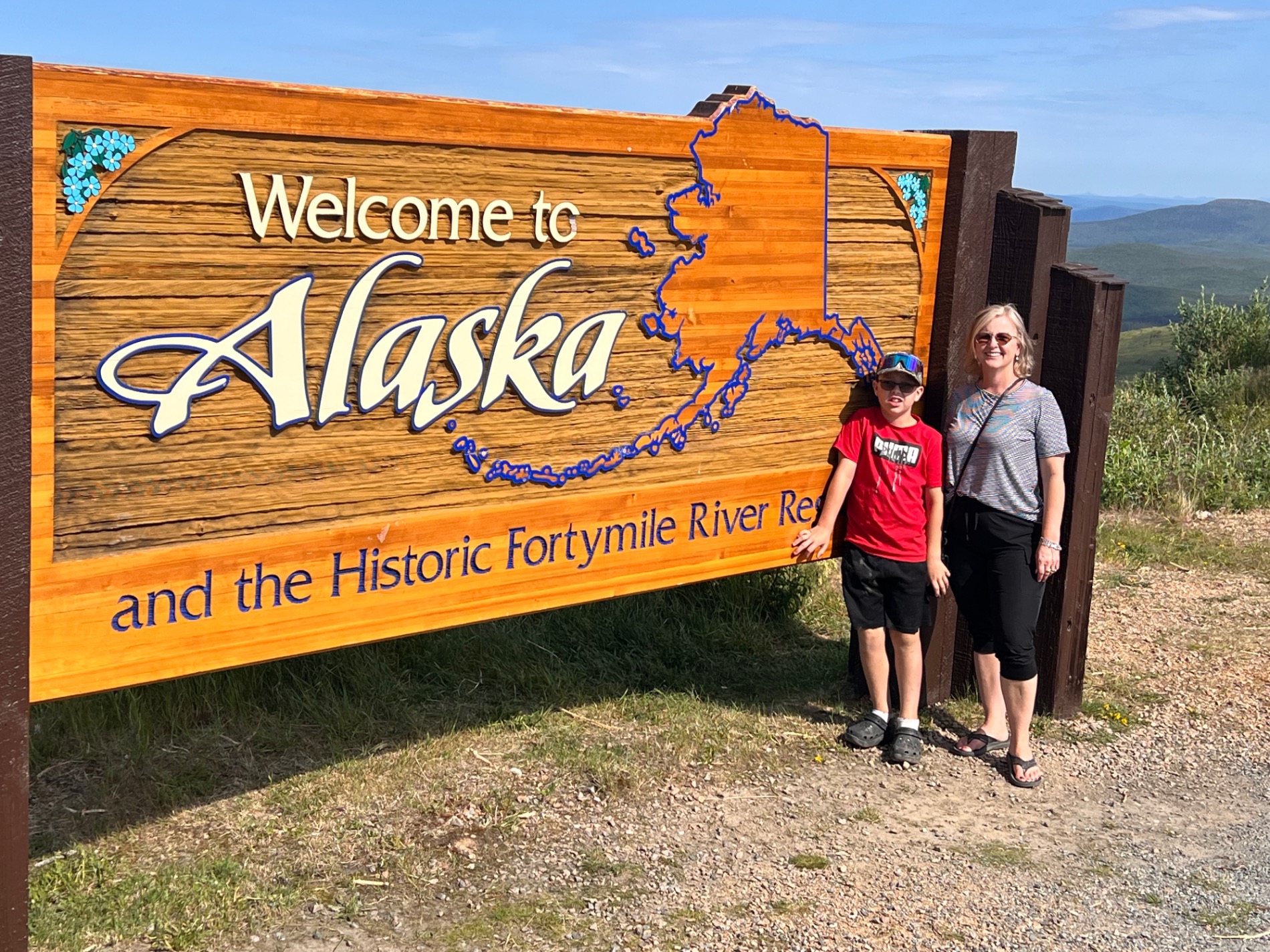 Two people posing at Welcome to Alaska sign featuring state map and Historic Fortymile River