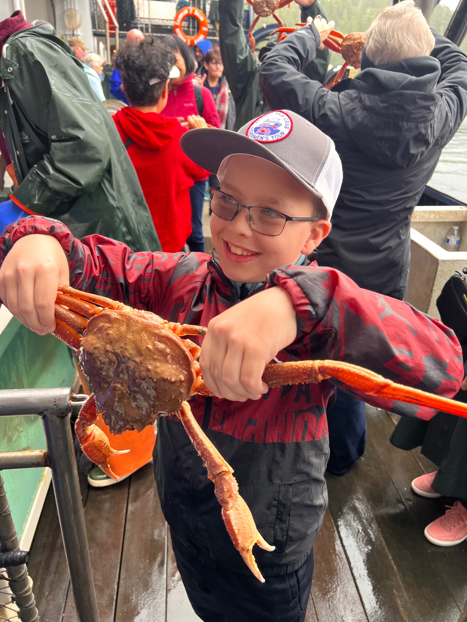 Young boy holding large red king crab on fishing boat deck with mountains behind