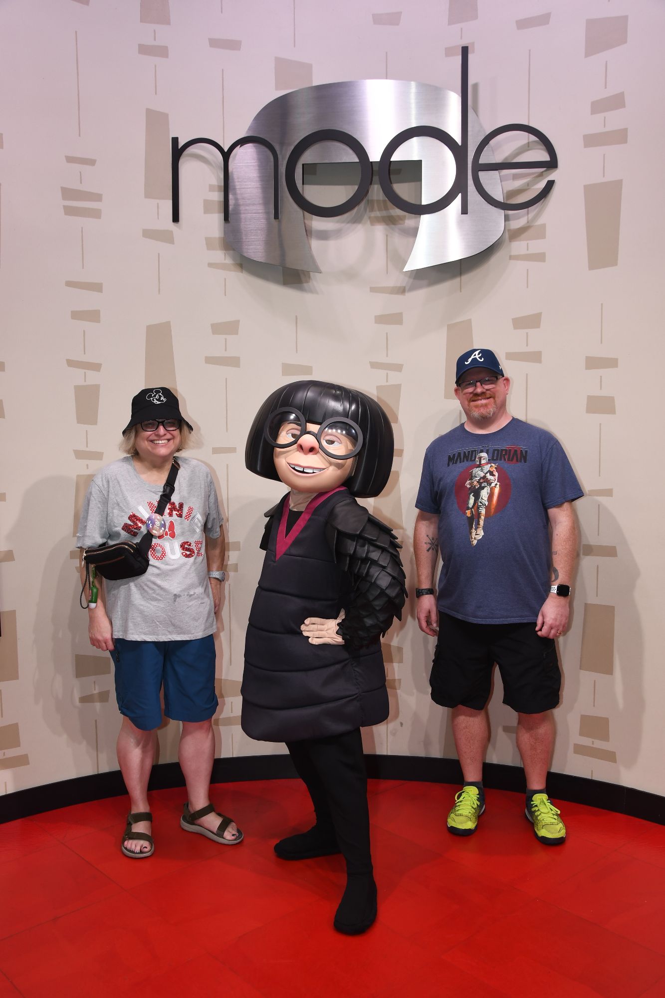 Two visitors and a character dressed as Edna Mode stand together on a red carpet in front of the Pixar 'mode' logo display