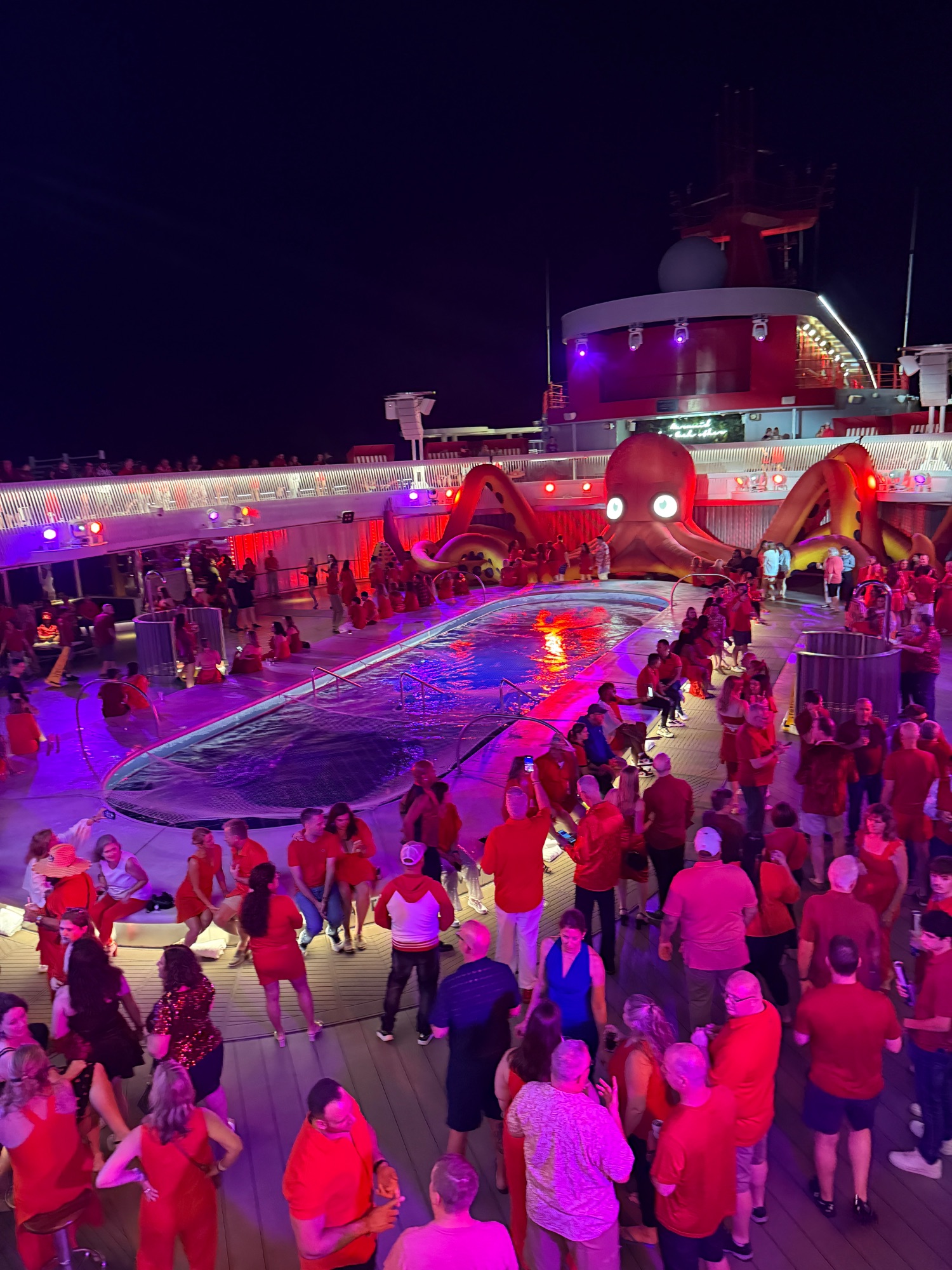 Nighttime pool deck party with colorful lighting, inflatable attractions, and crowds of passengers