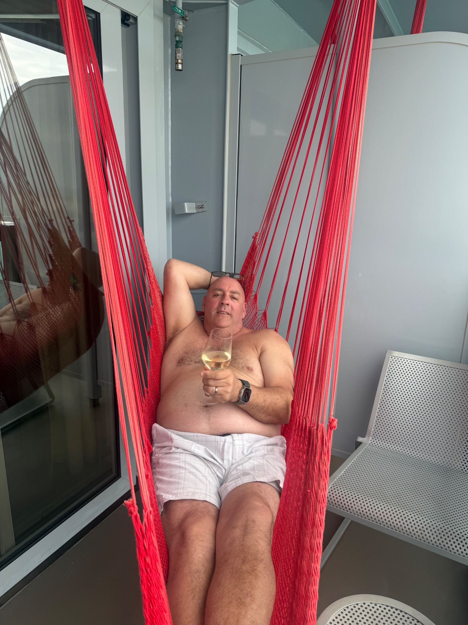 Man relaxing in red hammock on ship cabin balcony holding white wine glass