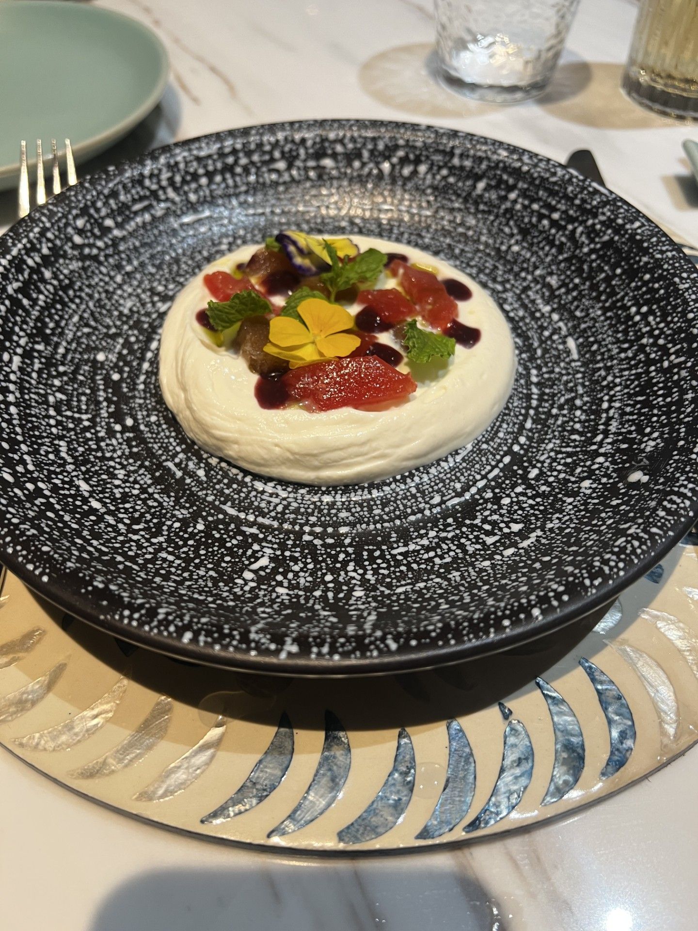 Minimalist plated dessert with cream, red fruit coulis, and edible flowers on a textured black plate