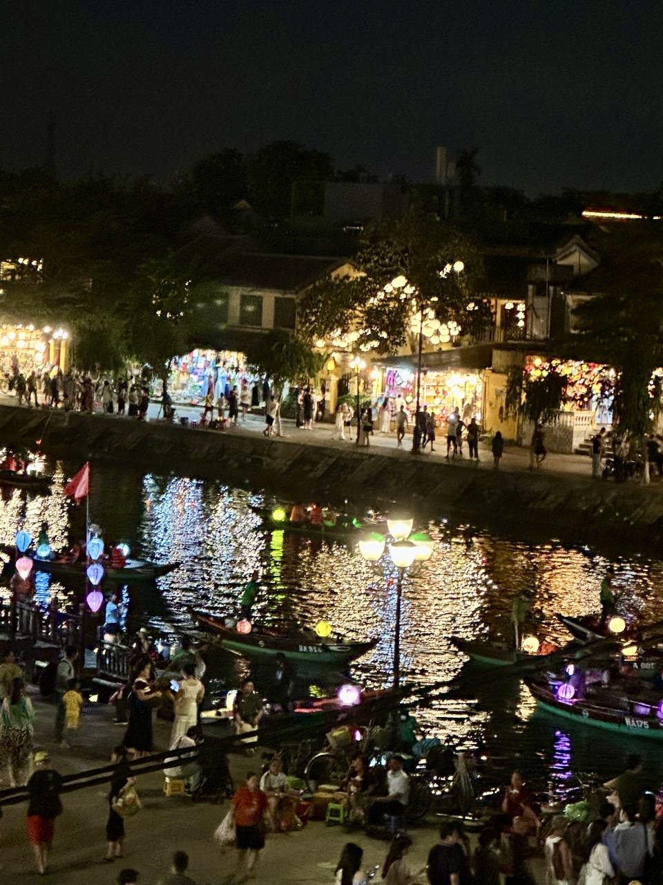 Hoi An riverside at night with colorful lantern-lit boats and crowds along the waterfront promenade