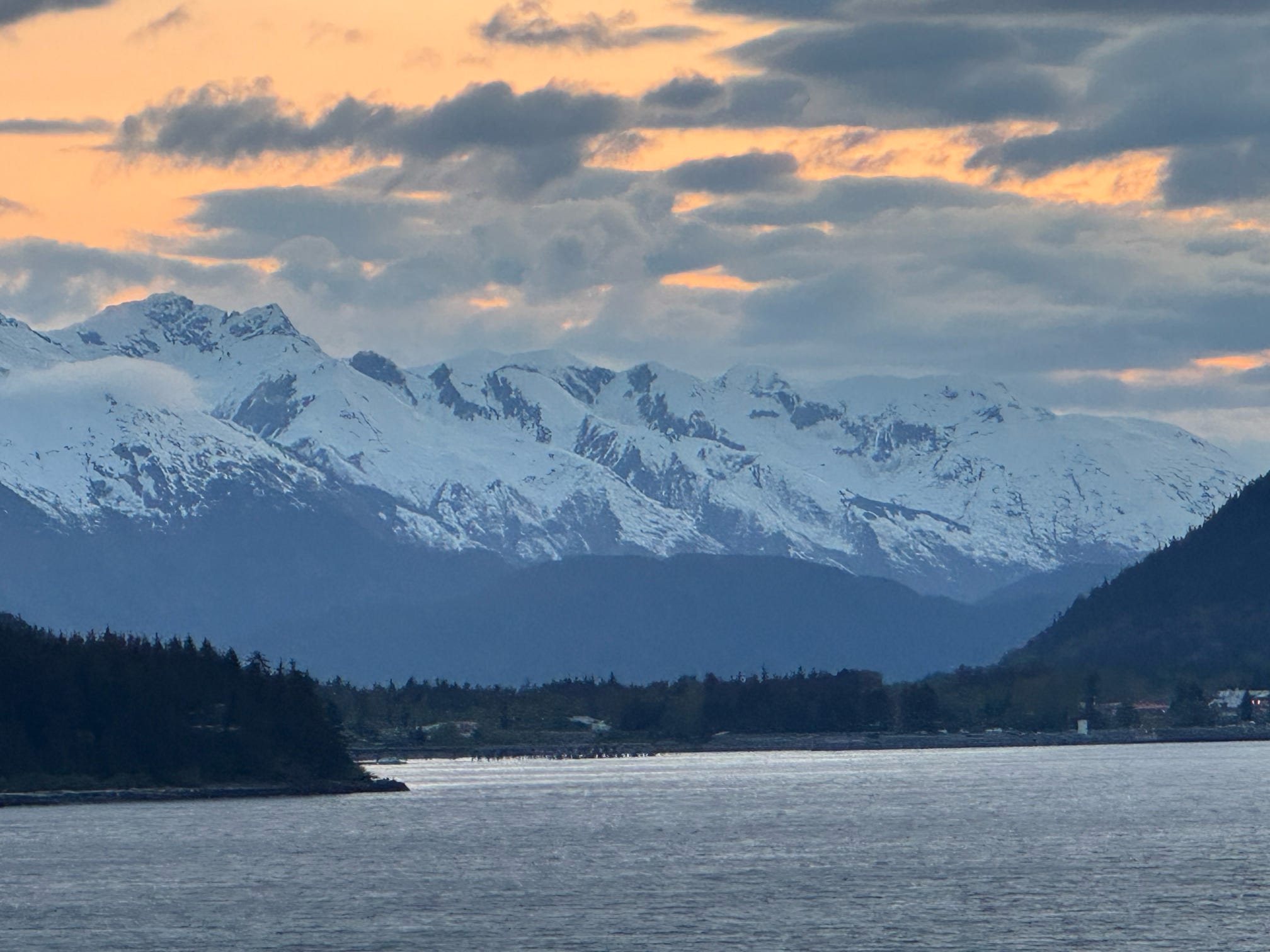 Snow-capped Alaskan peaks glow under a dramatic orange sunset sky reflected in calm coastal waters.
