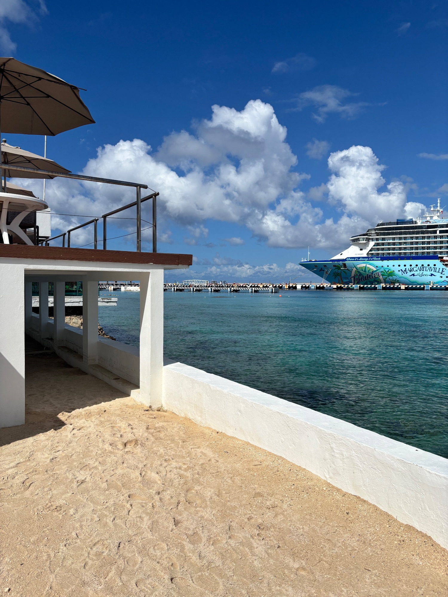 Beach view of Margaritaville cruise ship docked at Cozumel pier with white sand, turquoise water, and blue sky with white clouds