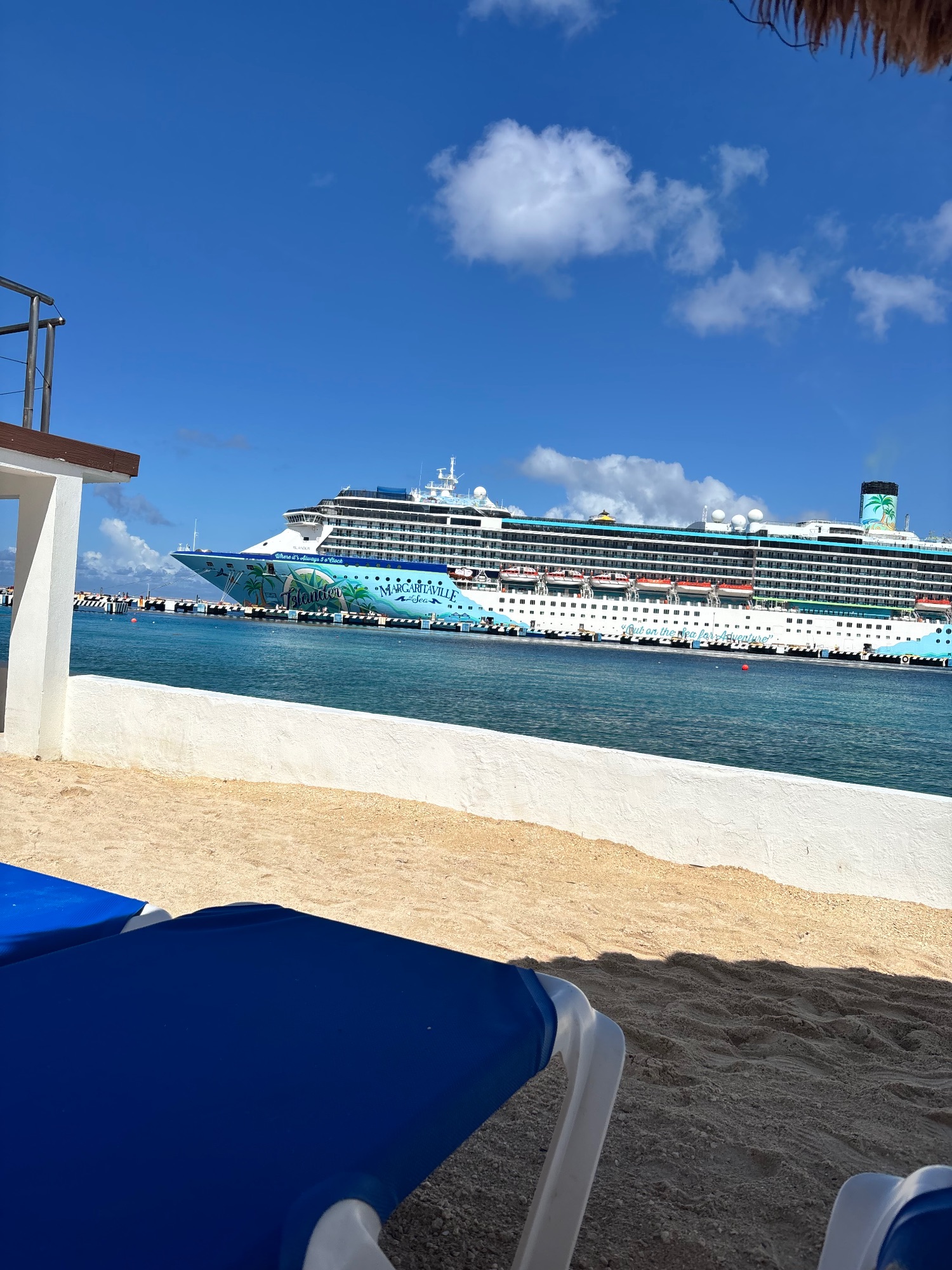 Margaritaville cruise ship moored at Cozumel dock viewed from sandy beach with lounge chair in foreground and clear blue sky