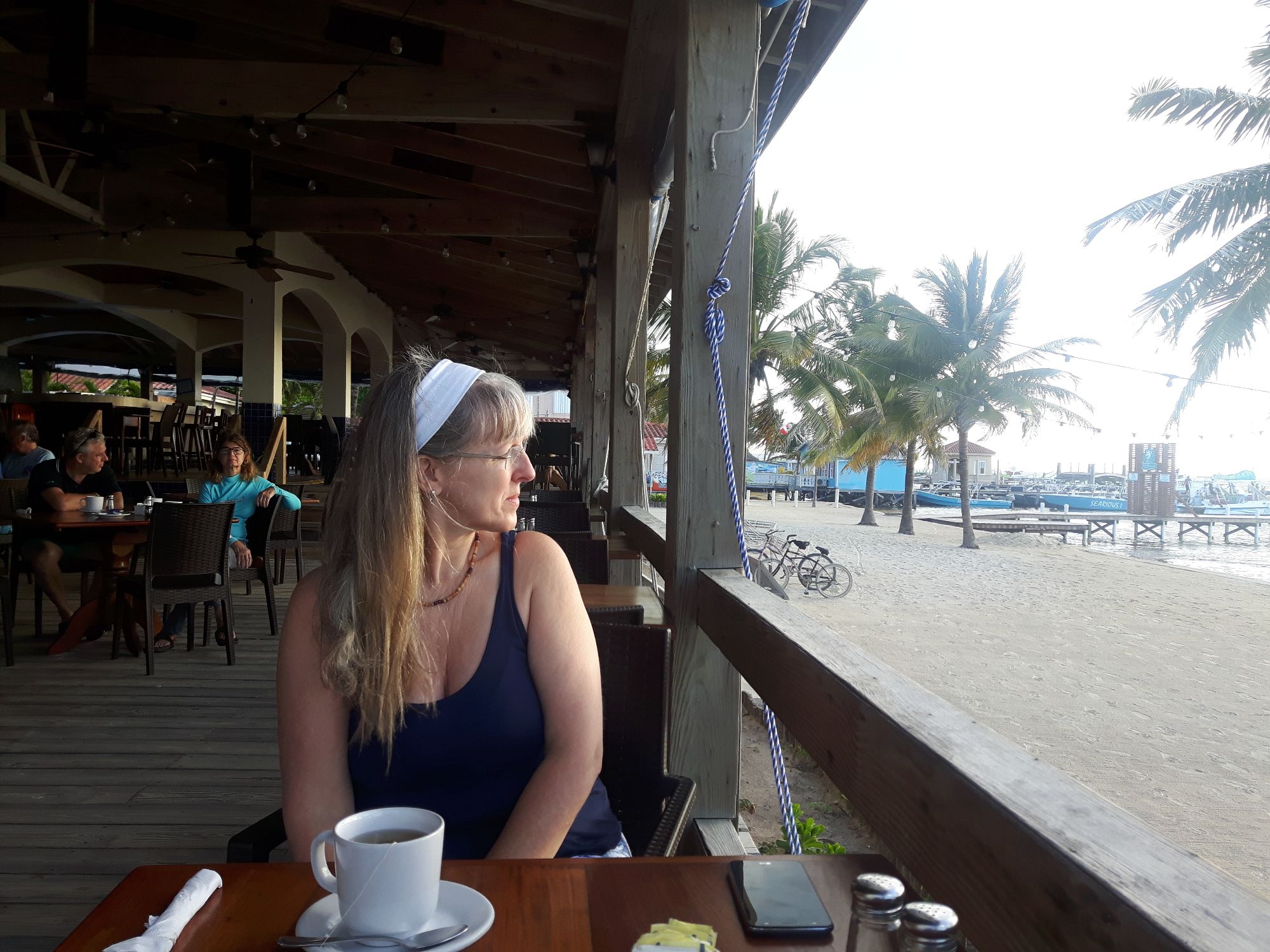 Woman in navy tank top sitting at a beachfront restaurant table with coffee, overlooking sand and palm trees