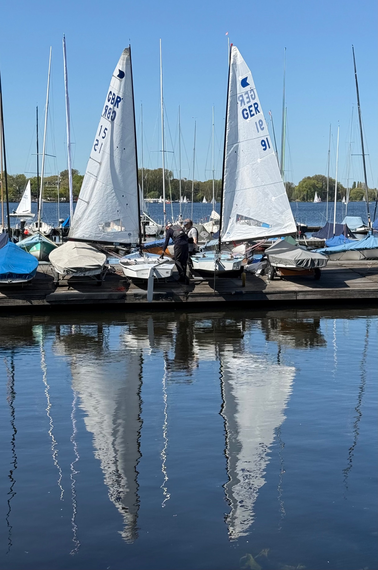 Two white sailing dinghies with German registration numbers moored at a dock, reflected in calm water