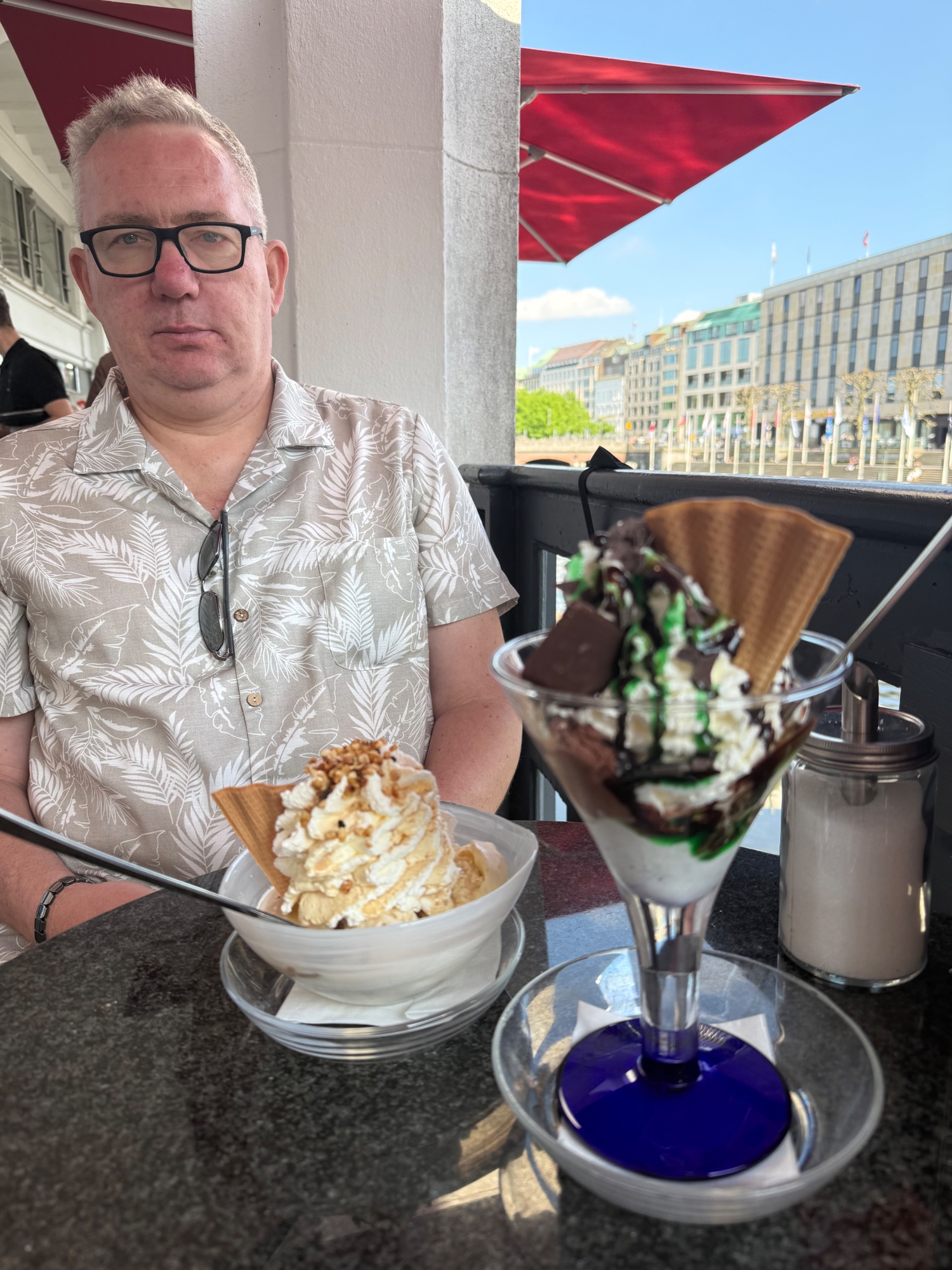 Man in patterned shirt holding a coffee with whipped cream at an outdoor café with red umbrella