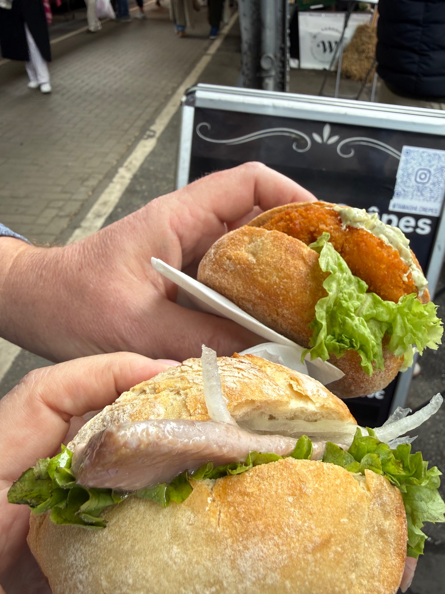 Hand holding a sandwich with sliced fish and lettuce on crusty bread at a street market stall