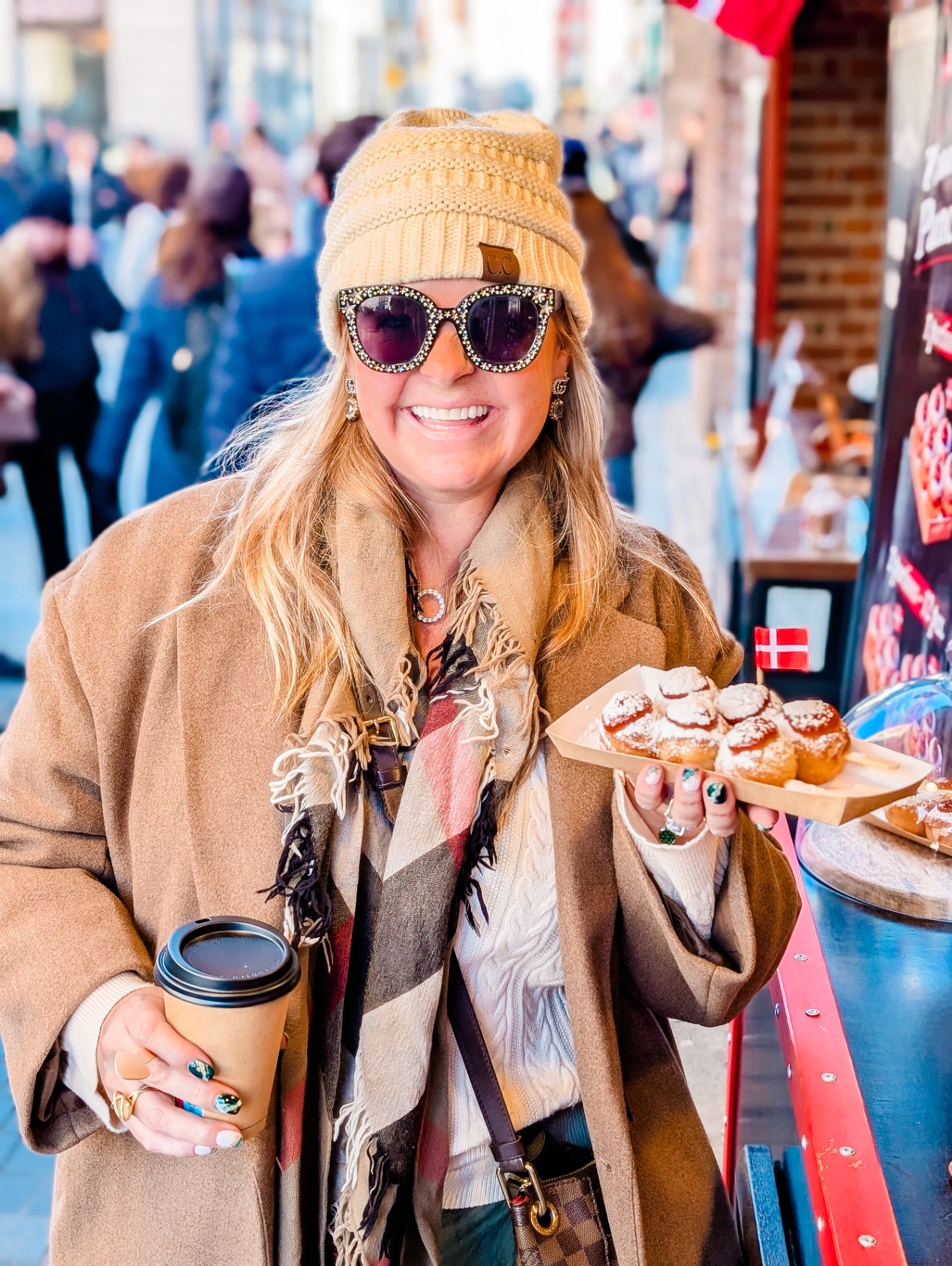 Woman in beige coat and white beanie holding coffee and pastry plate at an outdoor market with historic buildings in background