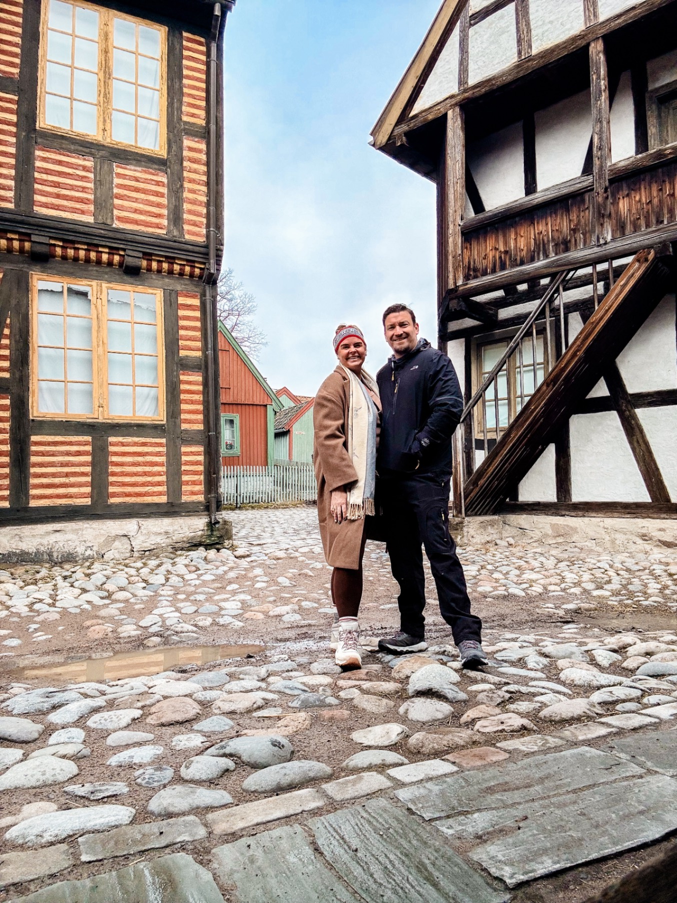 Two people standing between half-timbered medieval buildings with red and white striped wooden facades on a cobblestone street