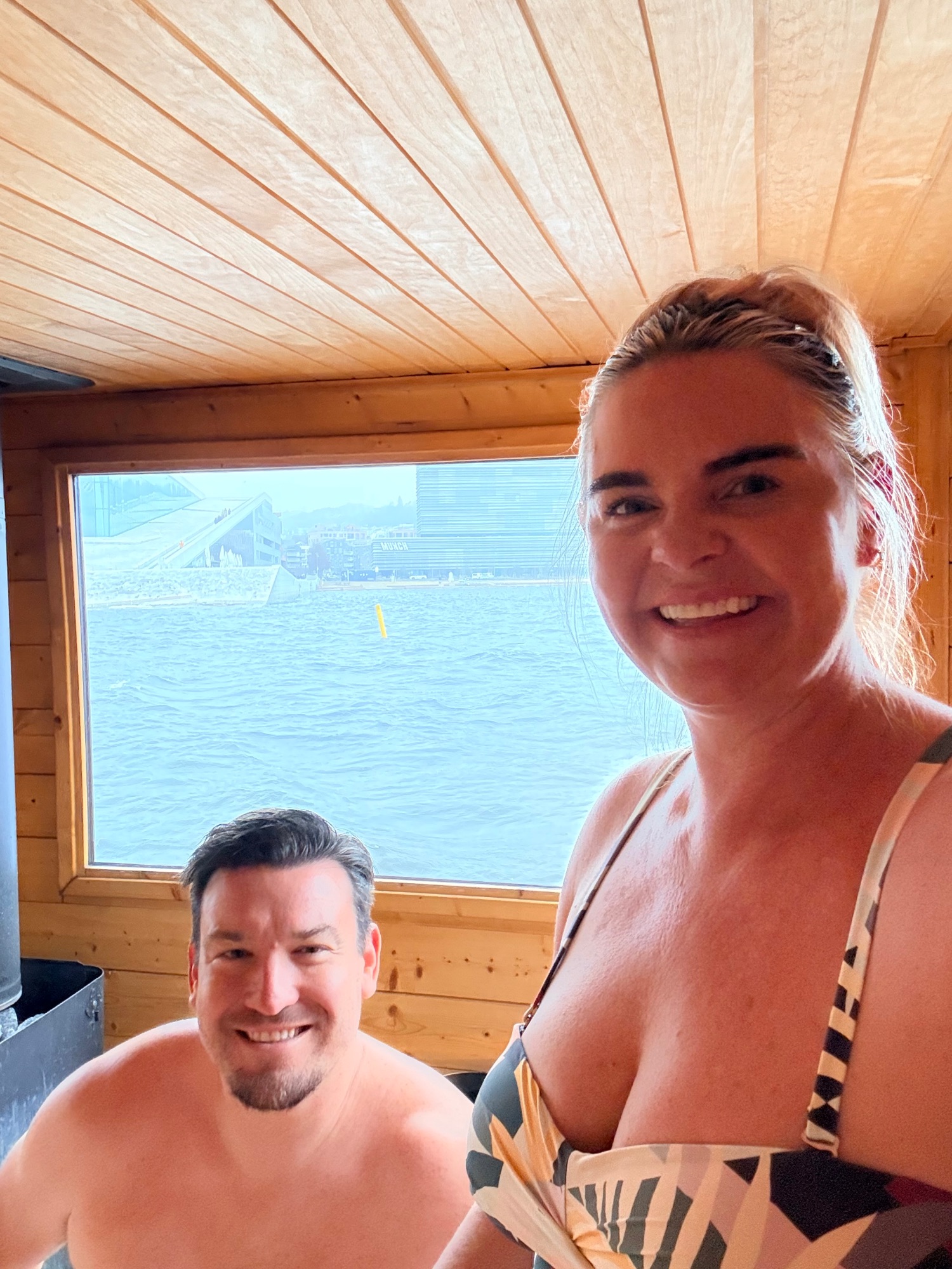 Two people relaxing in a wooden sauna or cabin interior with large window overlooking calm blue water