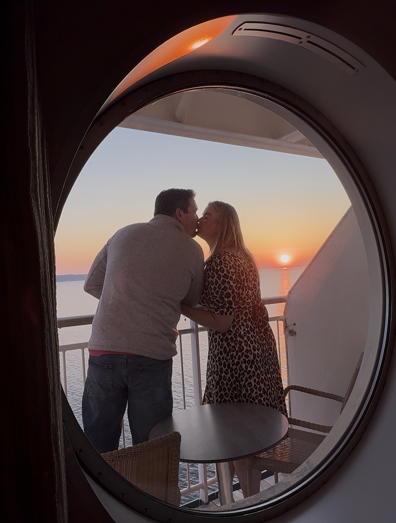 Couple kissing on a ship balcony at sunset with calm water and distant shoreline in background