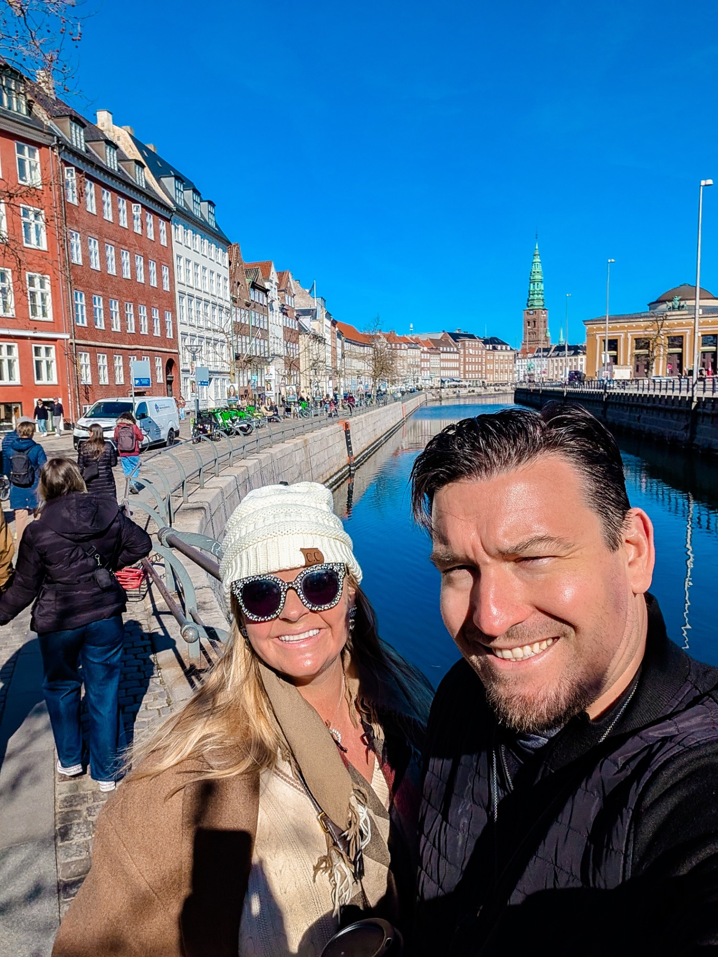 Two people smiling at camera with colorful historic waterfront buildings and boats reflected in calm water behind them in Copenhagen