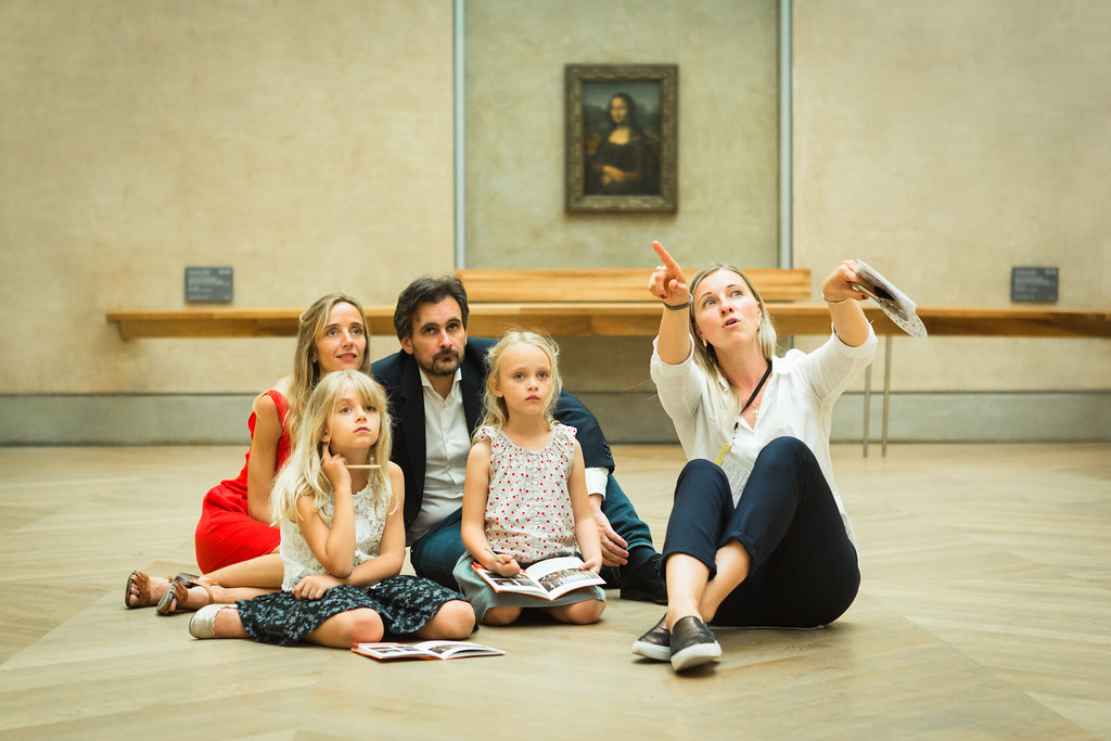 A woman and man sit with two young children on a wooden floor in front of the Mona Lisa at the Louvre, looking at printed materials