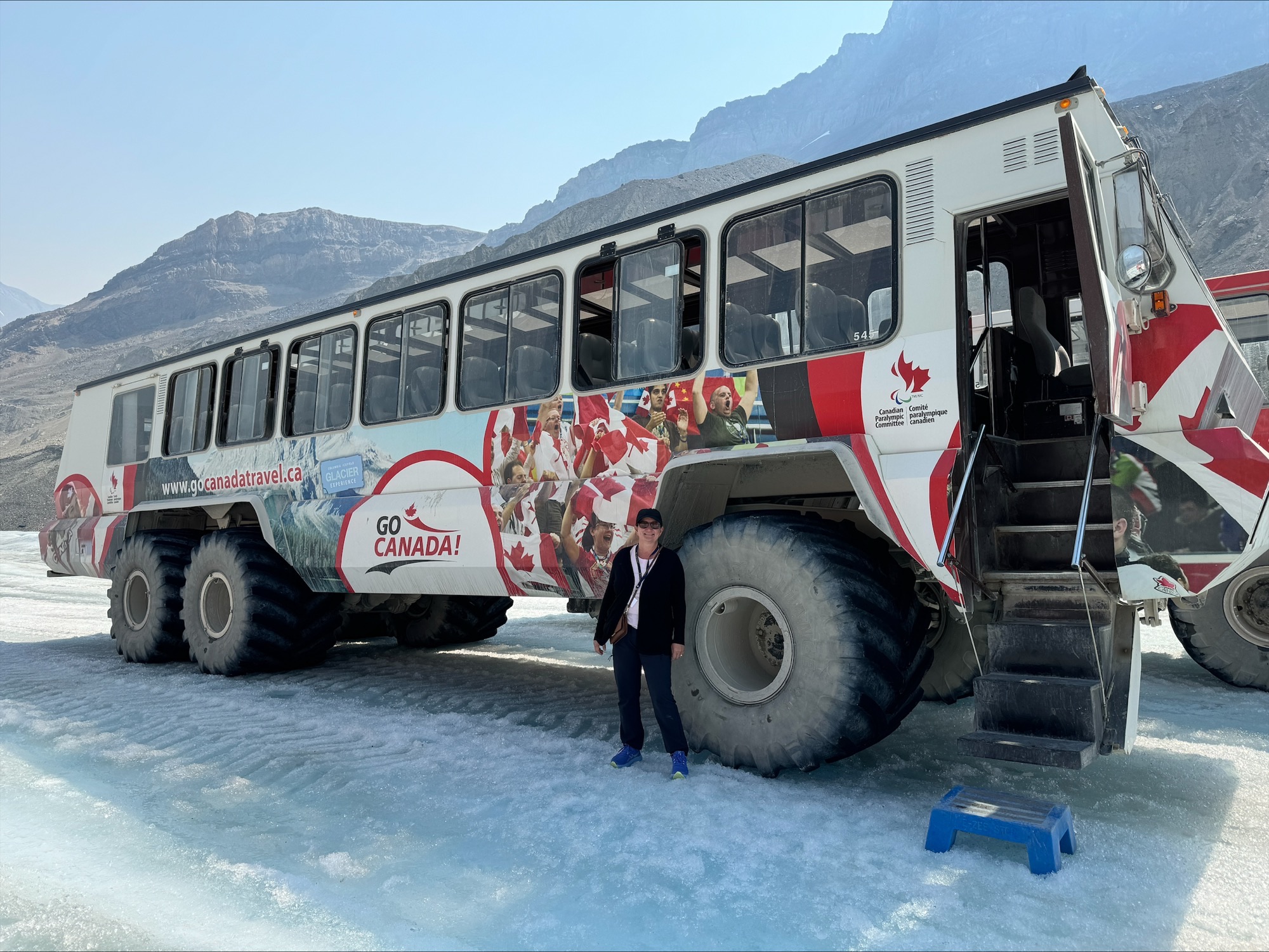 Donna standing in front of a red and white GO CANADA travel bus on a frozen lake with mountains in background