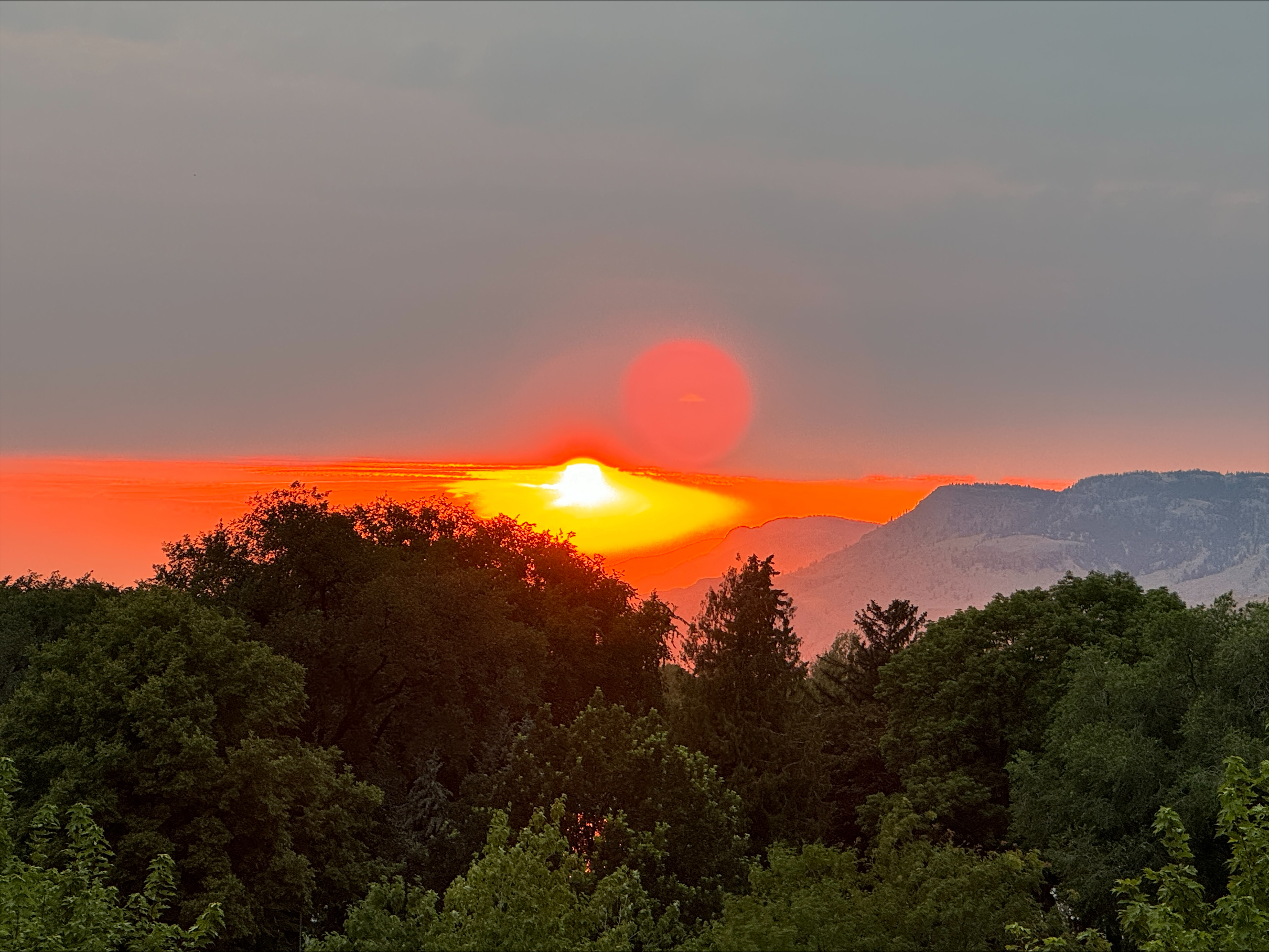 Vibrant sunset with large orange sun setting behind forested mountains and hills