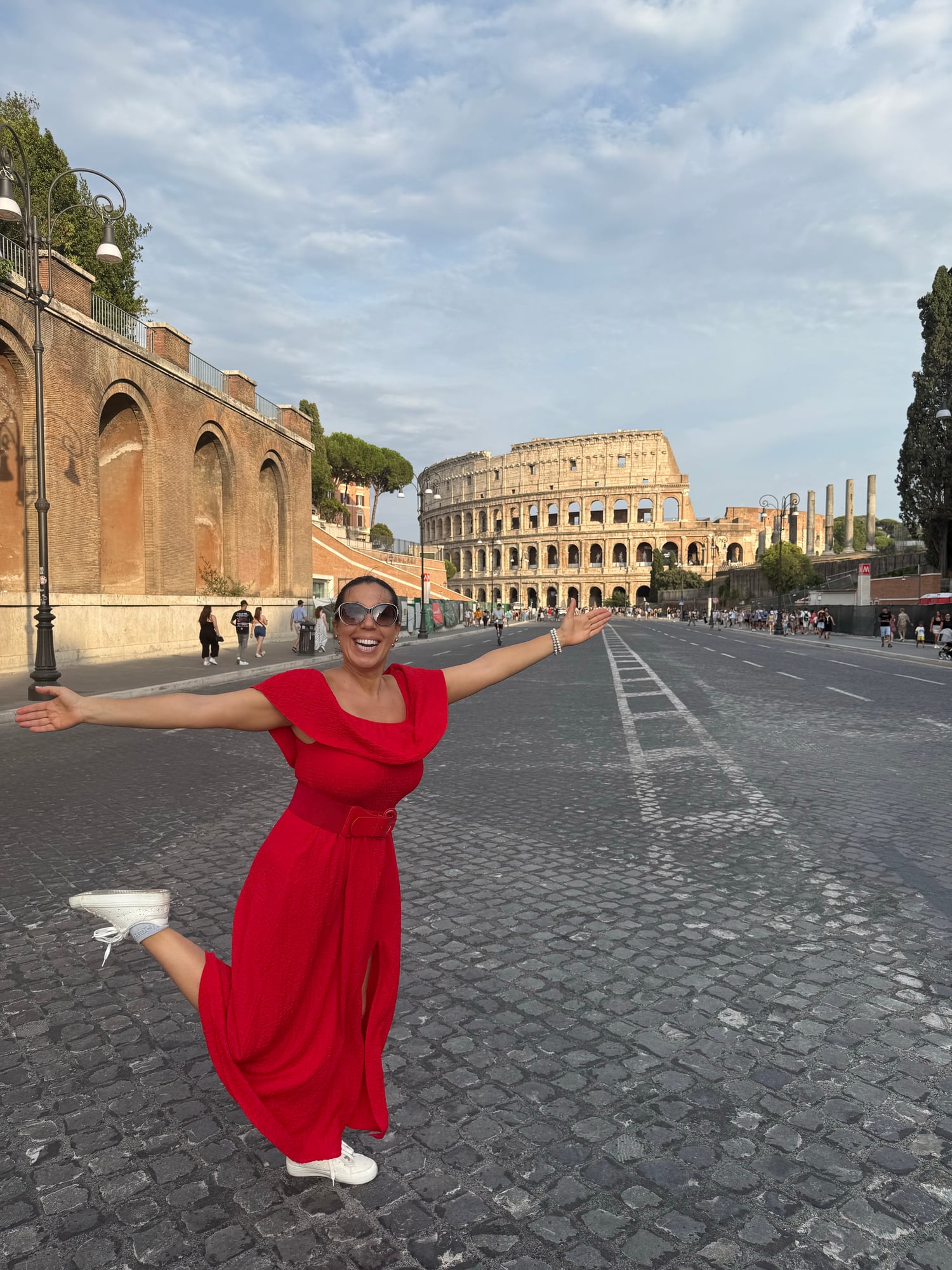 Katia stands with arms outstretched in front of the Colosseum in Rome, wearing a red dress and sunglasses