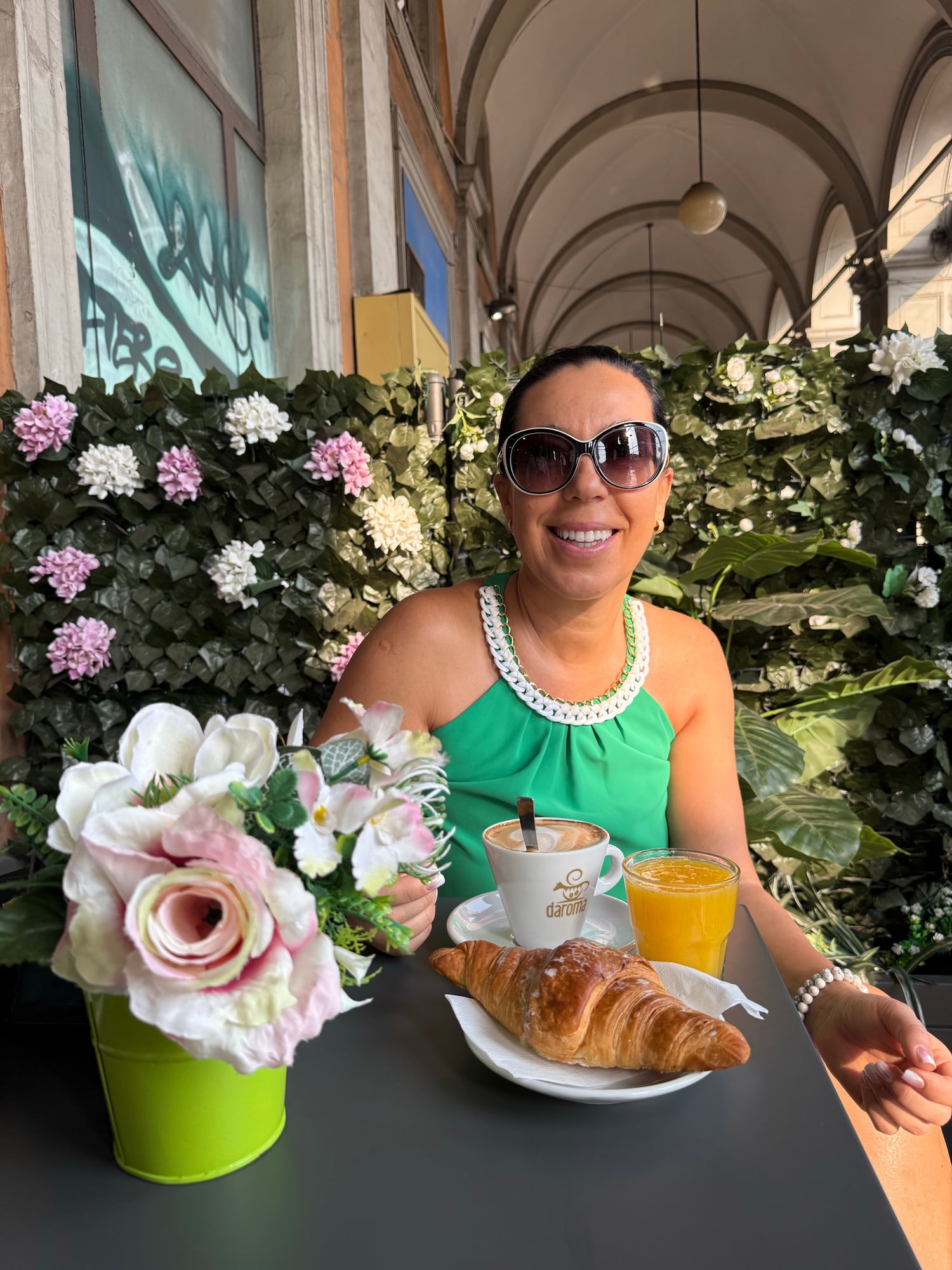 Katia smiles at a café table with a cappuccino, croissant, and orange juice, surrounded by pink and white flowers