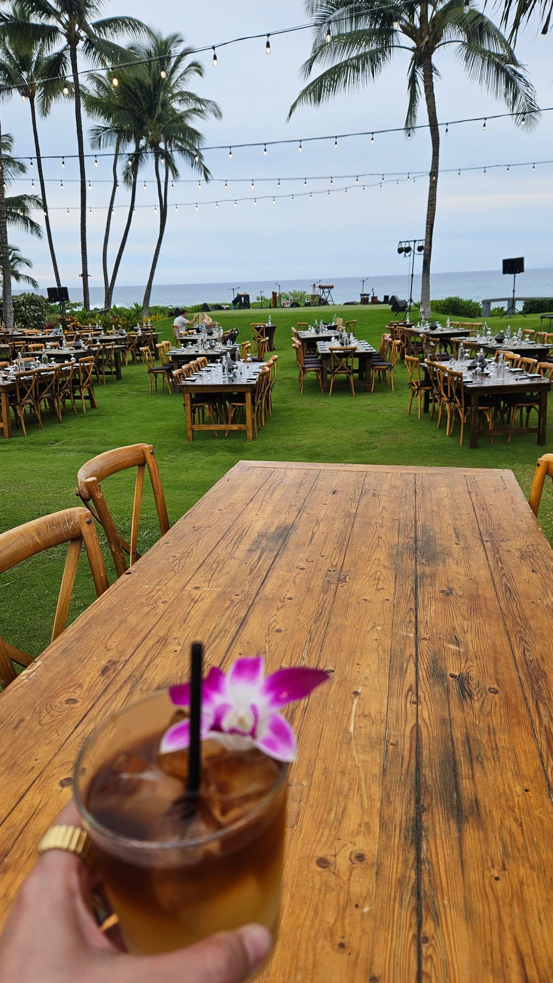 Oceanfront lawn dining setup with wooden tables, palm trees, string lights, and island views at a Maui resort
