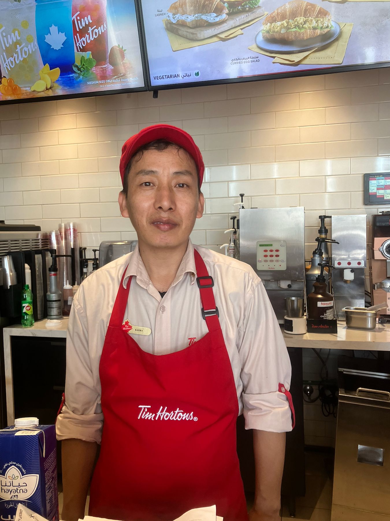 Tim Hortons employee in red uniform and cap stands behind counter with menu boards visible in background