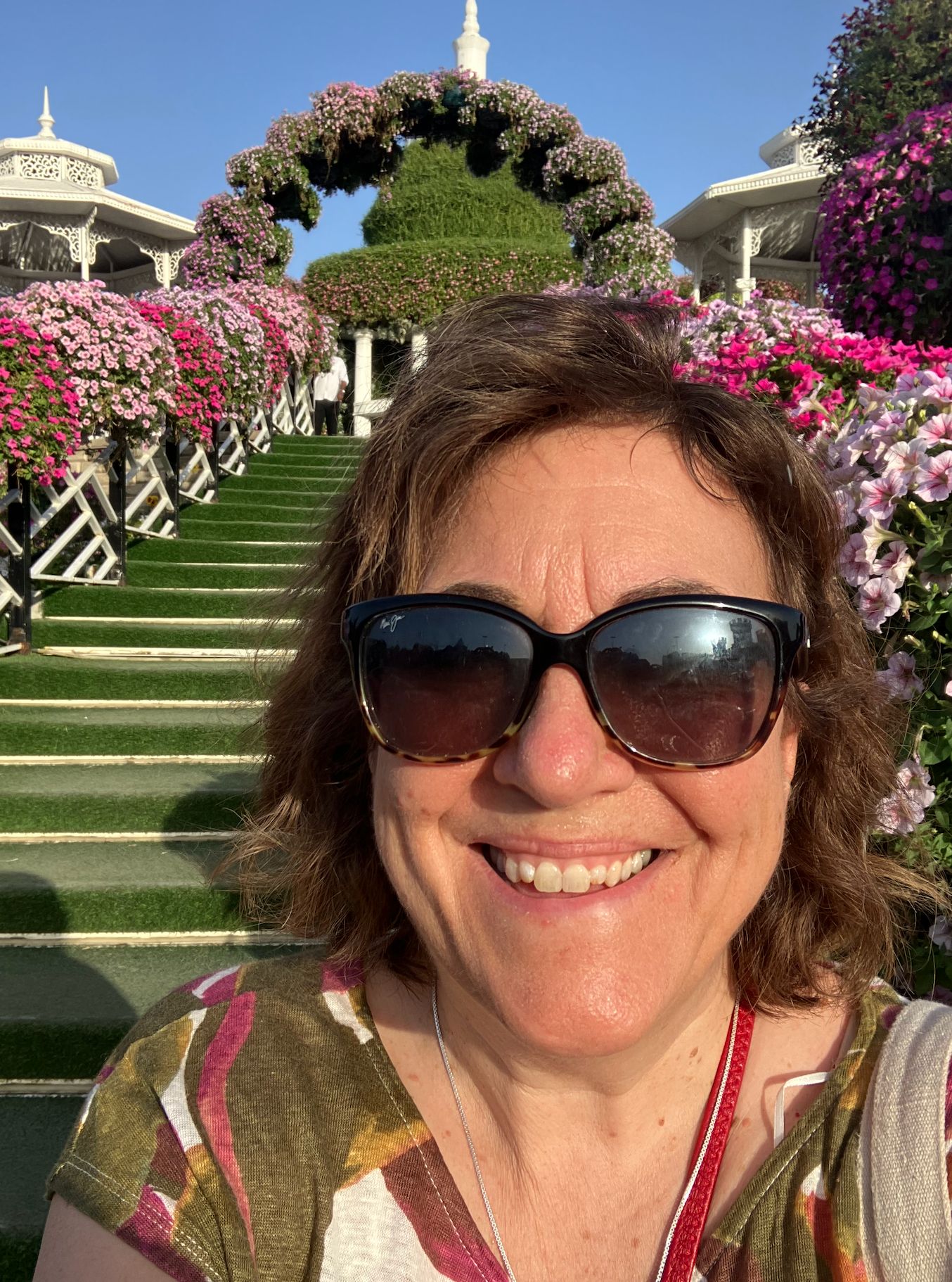 Woman in sunglasses smiles at camera with pink flower-lined staircase and white structures behind her