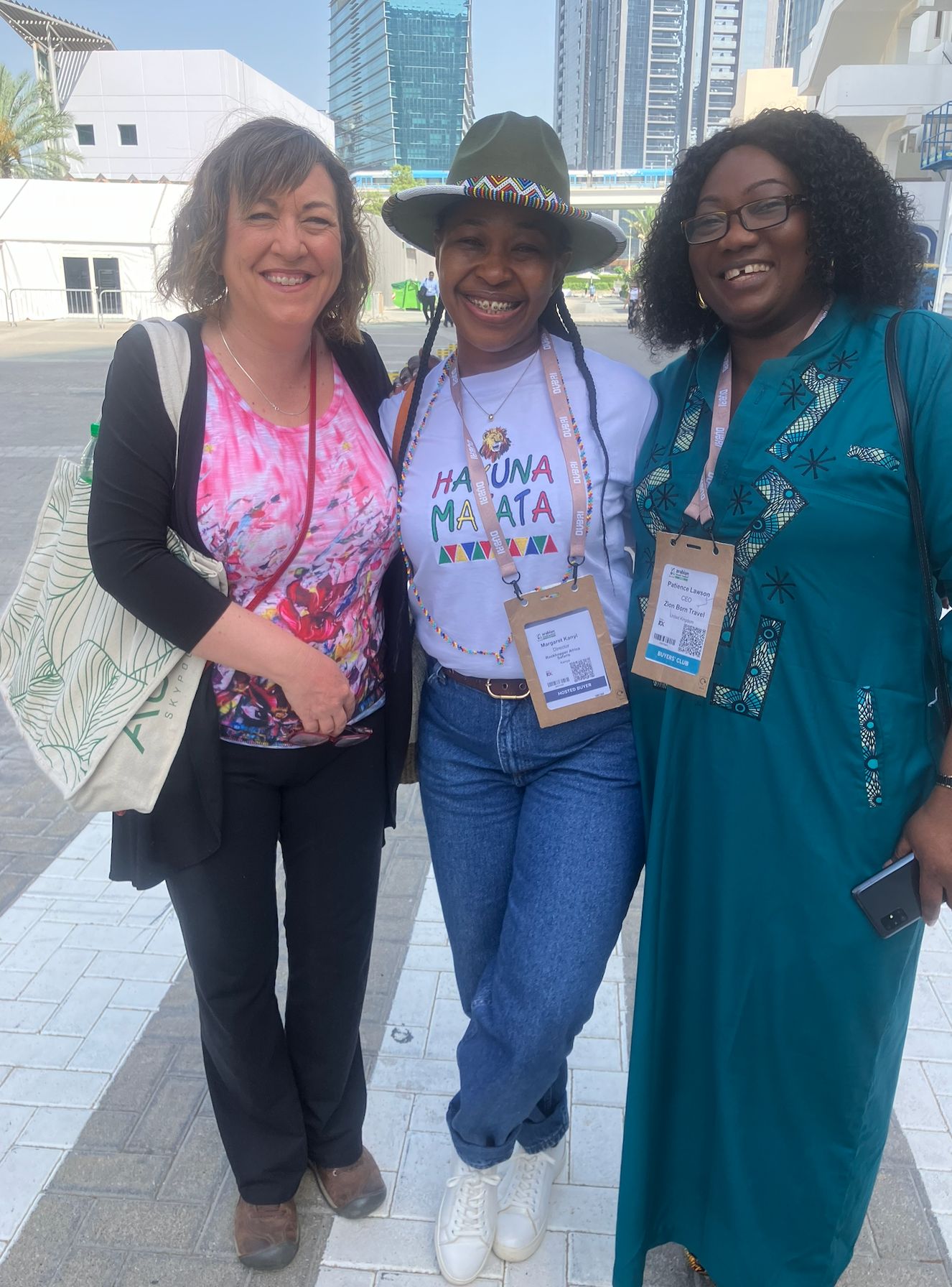 Three women wearing event lanyards stand together outdoors with modern buildings visible in background