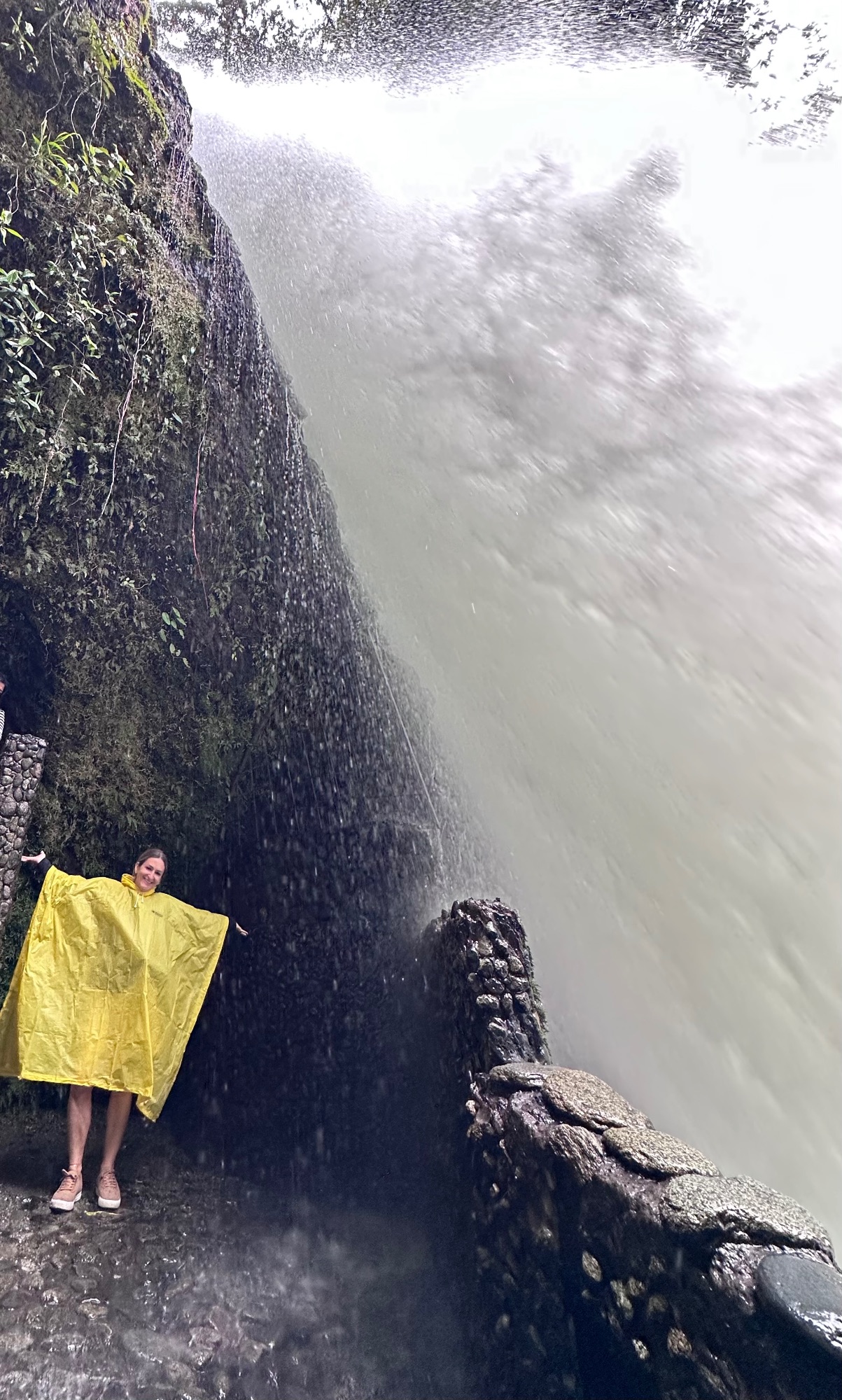 Person in yellow raincoat standing on wet rocks beside a tall waterfall cascading down a cliff face