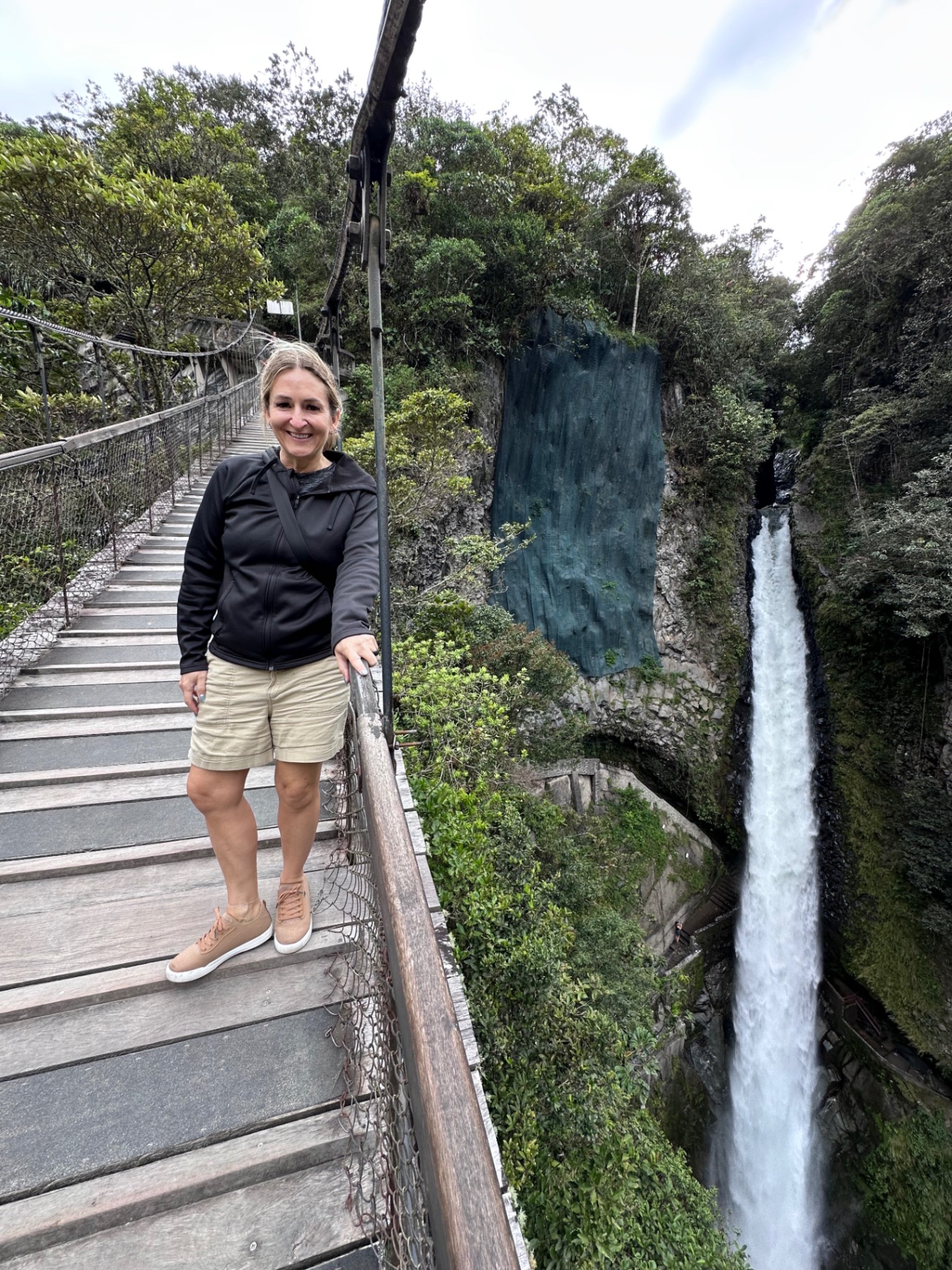 Woman smiling on a suspension bridge over a forested canyon with a waterfall visible in the background
