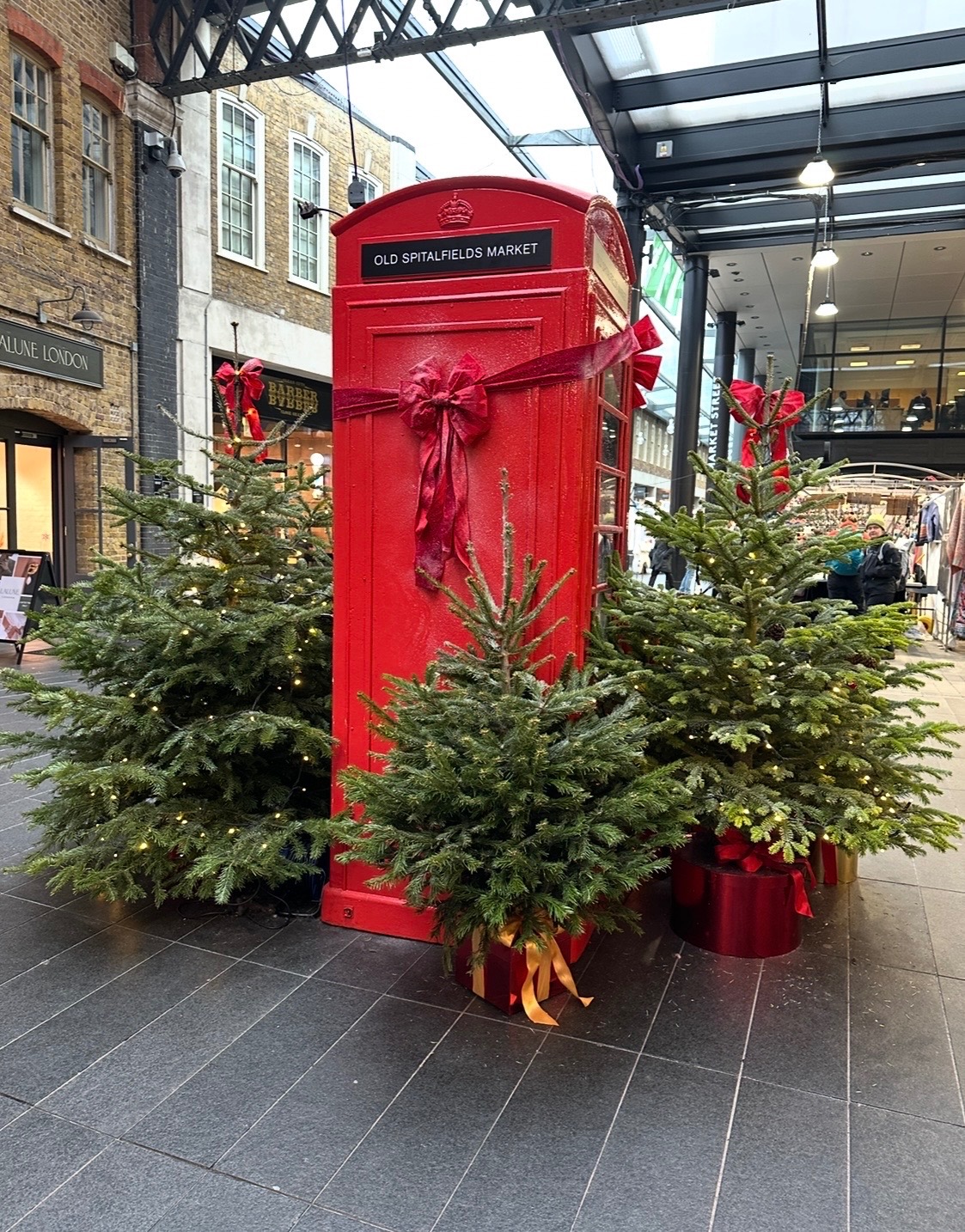 Red British telephone box decorated with evergreen garland and red ribbon bow, surrounded by small Christmas trees at Old Spitalfields Market