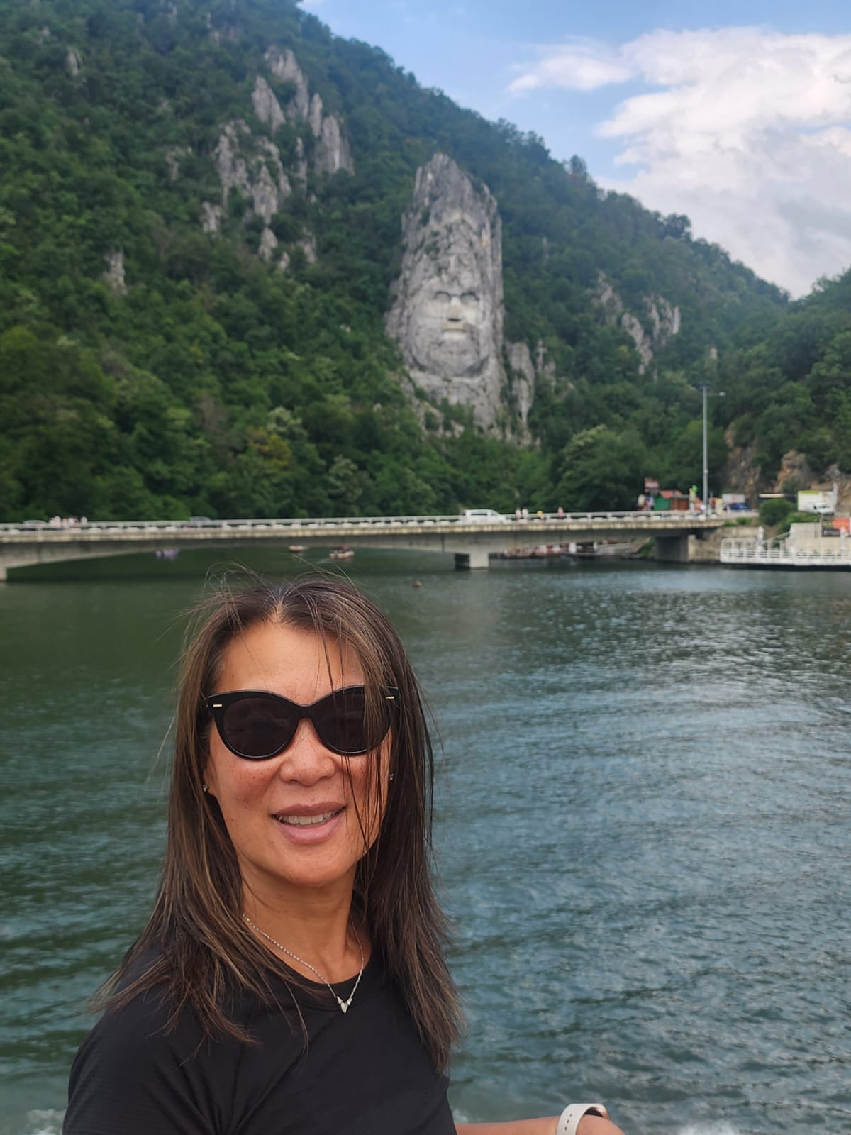 Grace Wong smiles at the camera with the Decebalus rock sculpture carved into a cliff face along the Danube River in Romania behind her