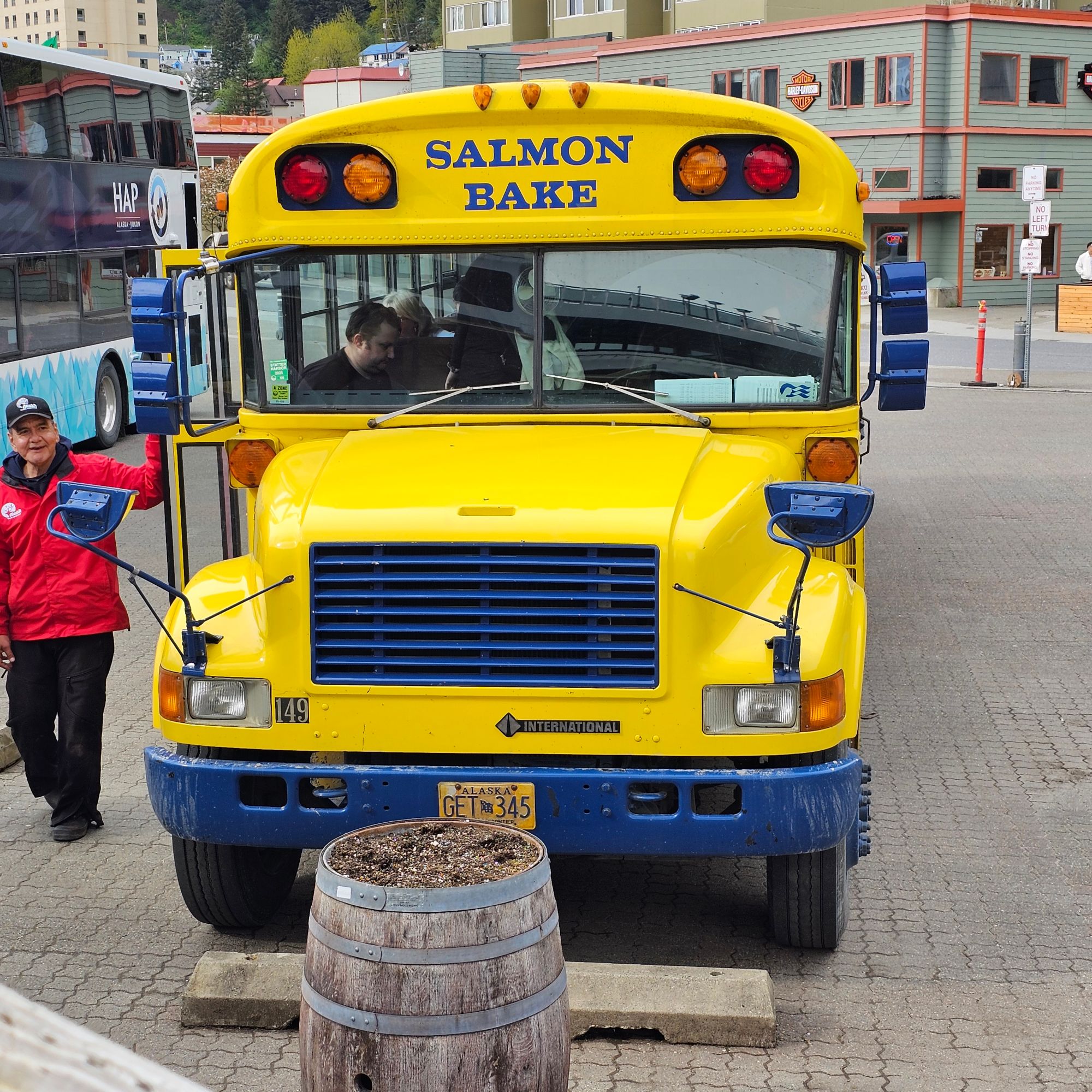 Bright yellow bus labeled 'Salmon Bake' parked on an Alaska street with a person standing beside it and a wooden barrel in front