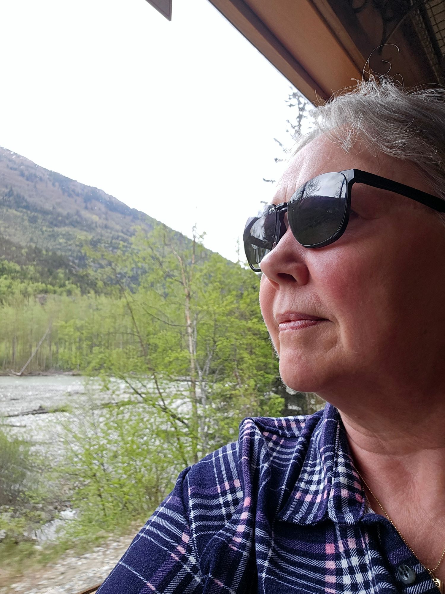 Woman with gray hair and sunglasses looking out from a boat toward a forested river valley with mountains in Alaska