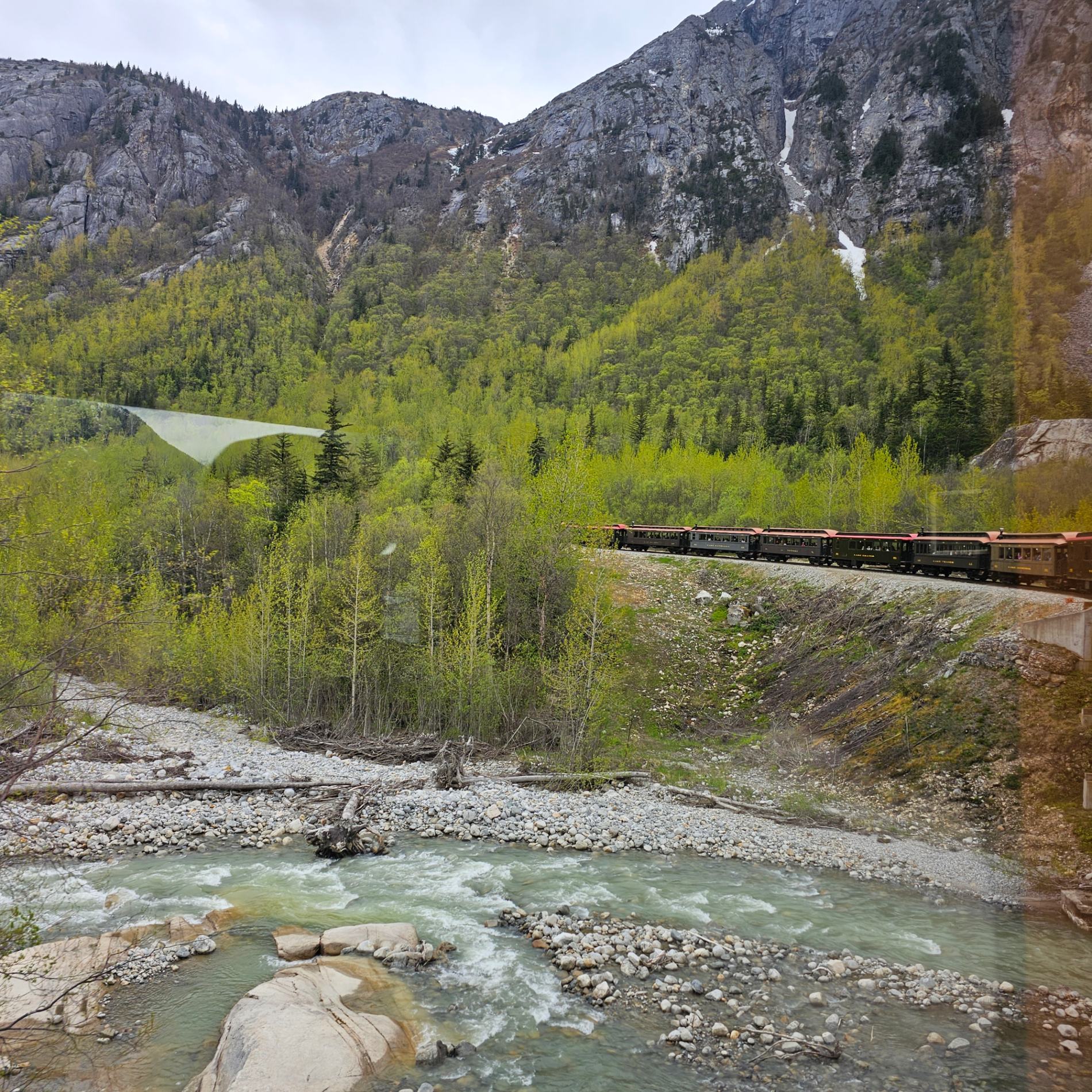 Scenic train traveling through a lush green valley with a turquoise river, rocky banks, and snow-capped mountains in the distance