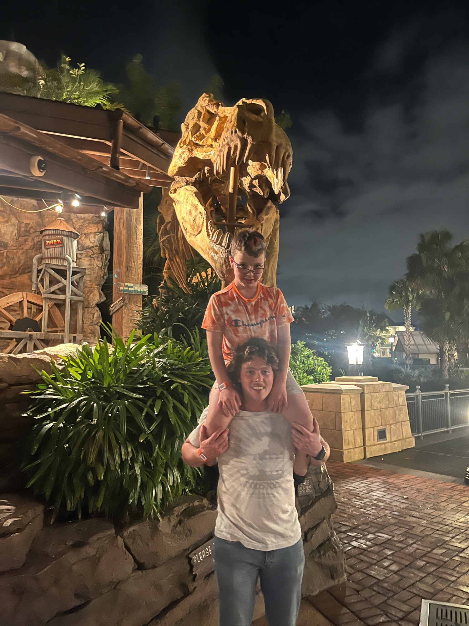 A young boy sits on an adult's shoulders in front of a large illuminated T-Rex dinosaur sculpture at night at a theme park