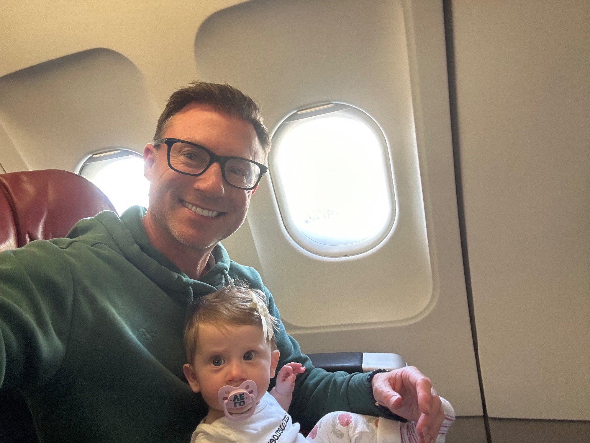 Man with glasses holding a baby on an airplane next to a window
