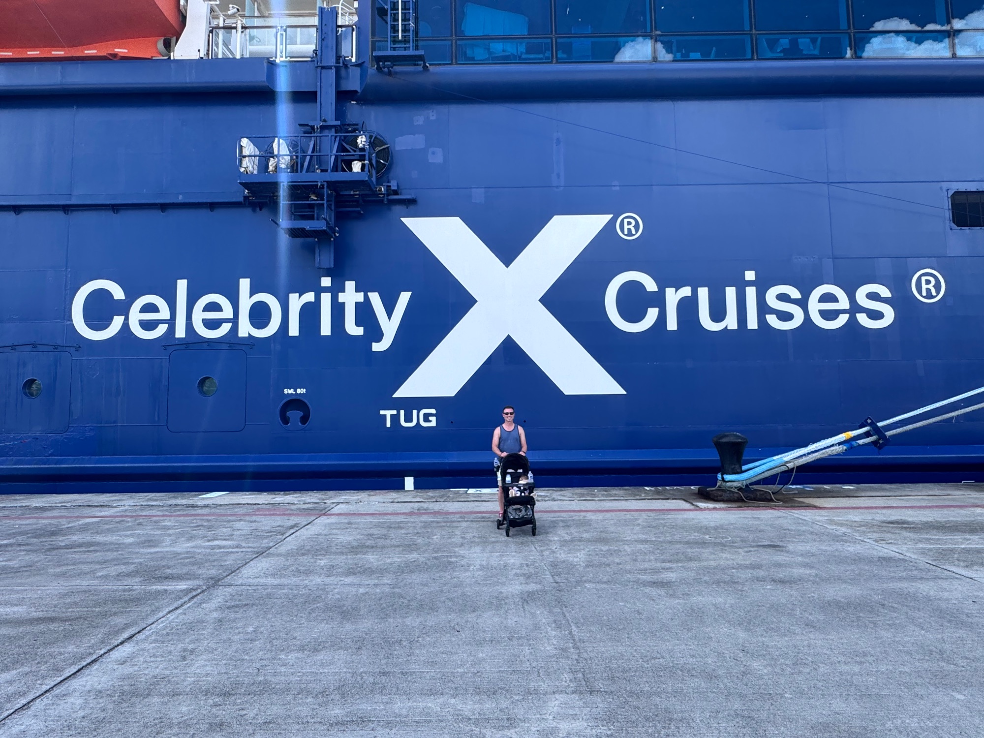 Man with a baby in a stroller in front of a large Celebrity Cruises ship hull