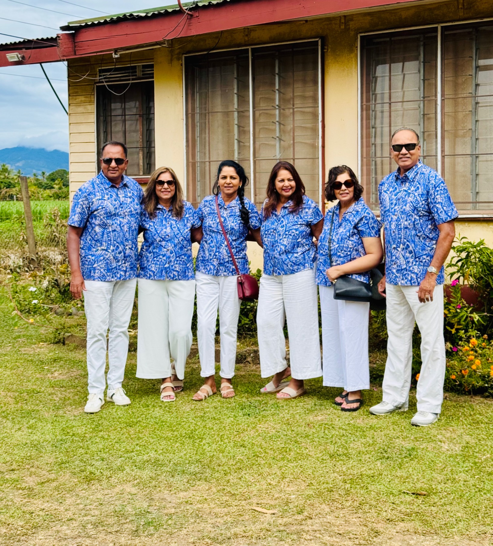 Six people in matching blue patterned shirts and white pants standing in front of a house with mountains