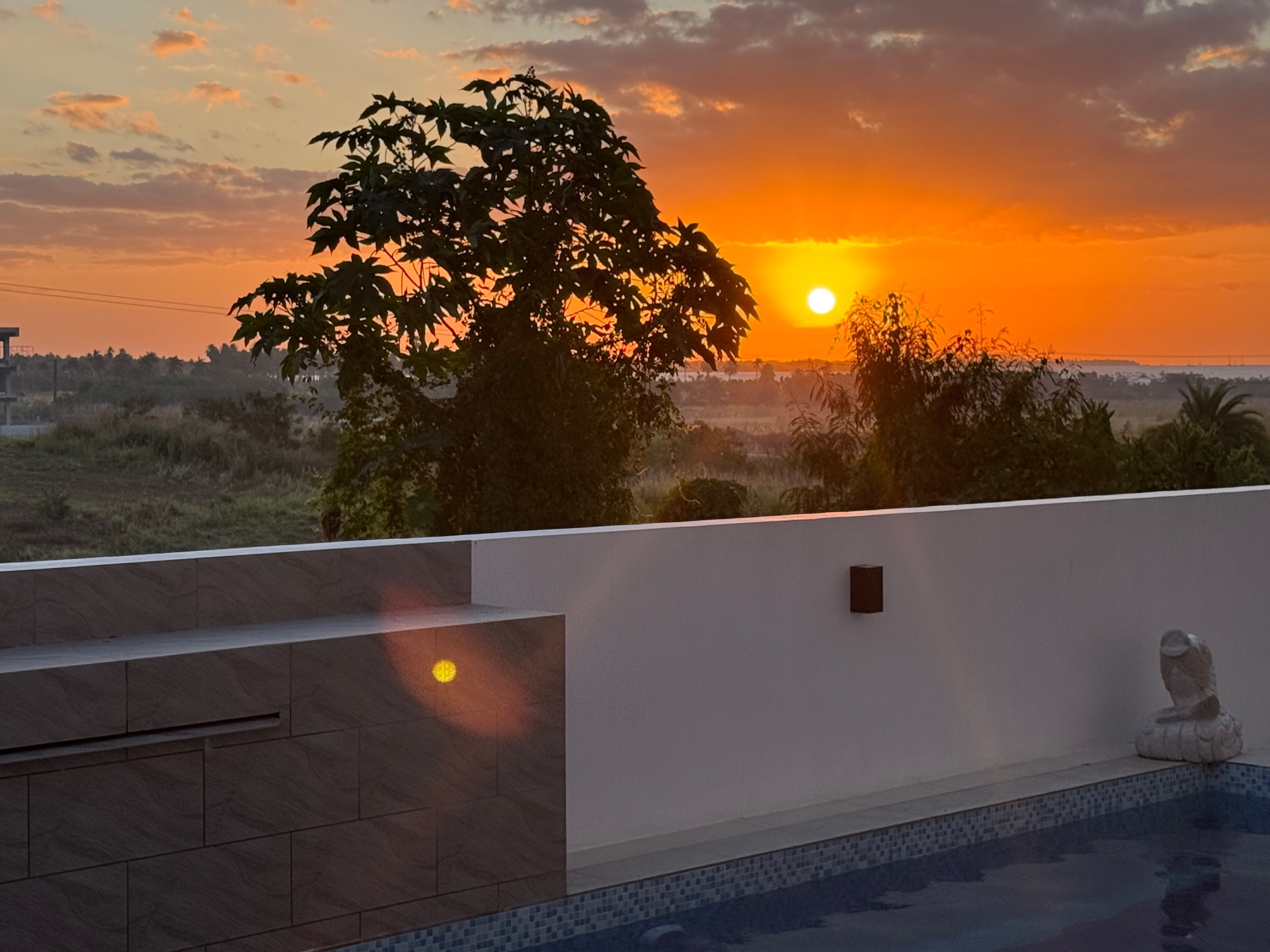 Sunset over rural landscape with silhouetted trees and modern white building with pool in foreground