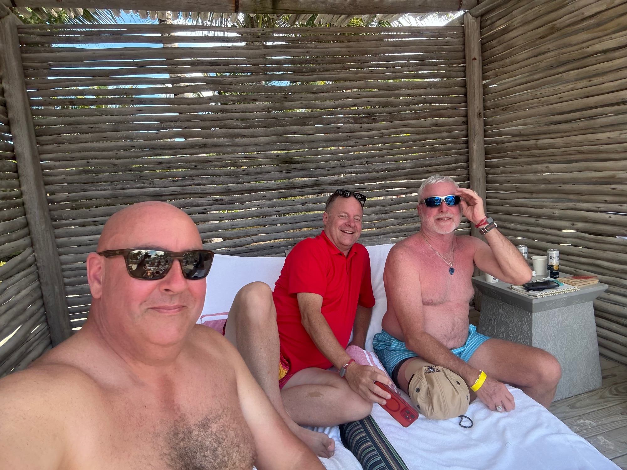 Three men in casual beach attire sitting together under a wooden pergola structure, smiling at the camera