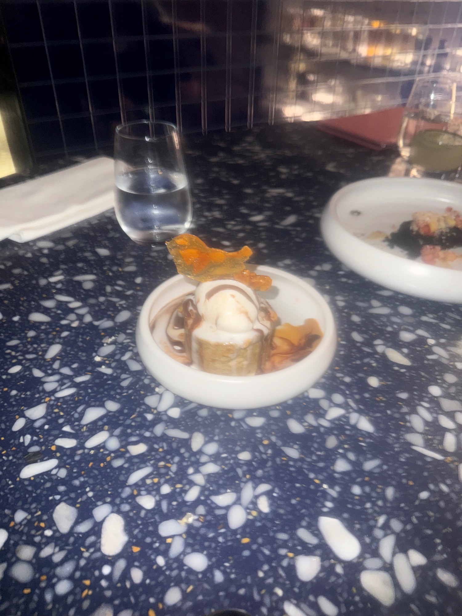 Plated dessert with vanilla ice cream and caramel decoration on a dark speckled table with water glass and dining setup