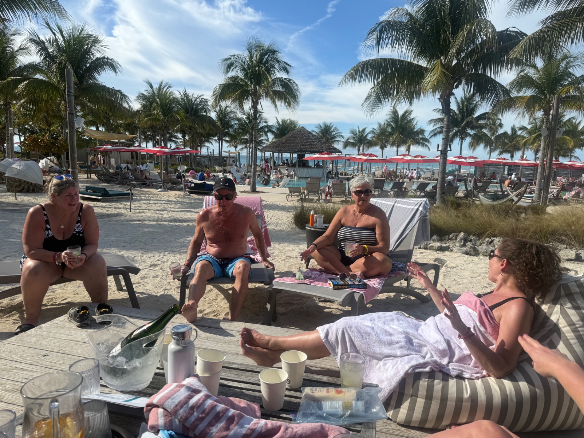 Group of people relaxing on beach loungers under red umbrellas with palm trees and ocean visible in background