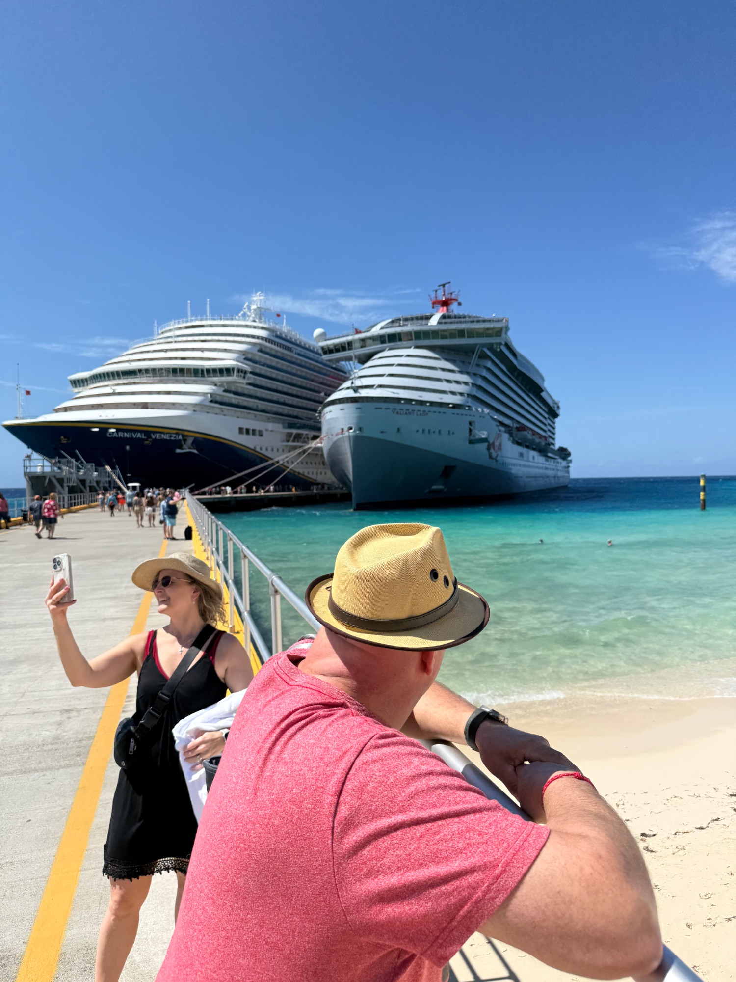 Two cruise ships docked at a Caribbean port with turquoise water, passengers on dock with cruise ship in background