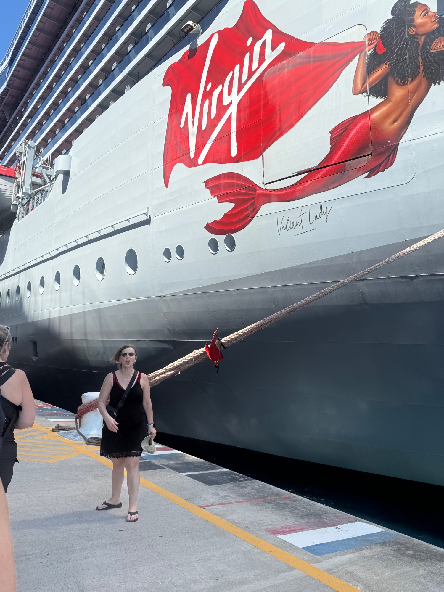 Woman in black dress standing at dock in front of Virgin Voyages cruise ship with red mermaid logo