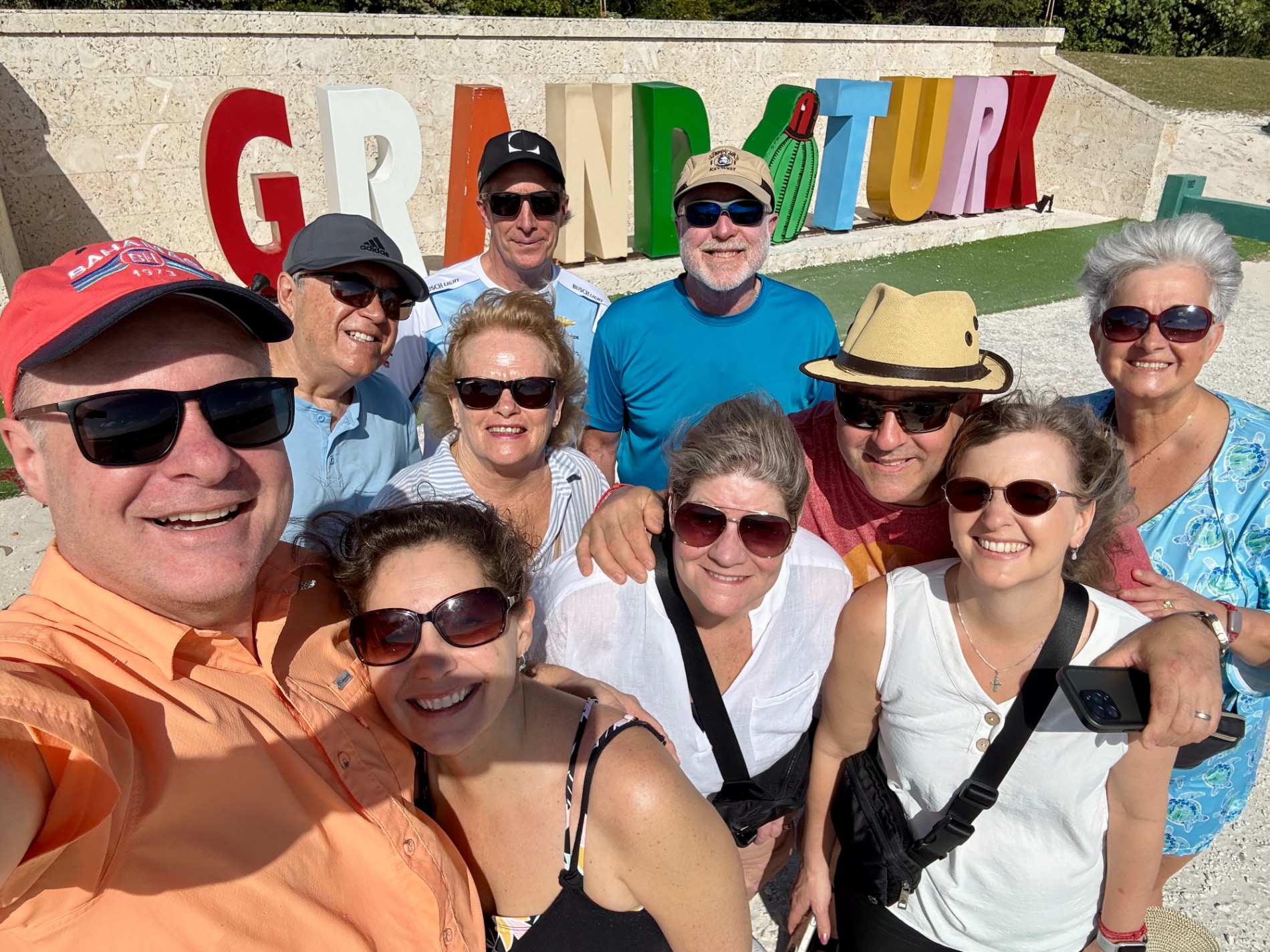 Group of eleven people posing together at GRAND TURK sign location, wearing sunglasses and casual vacation attire